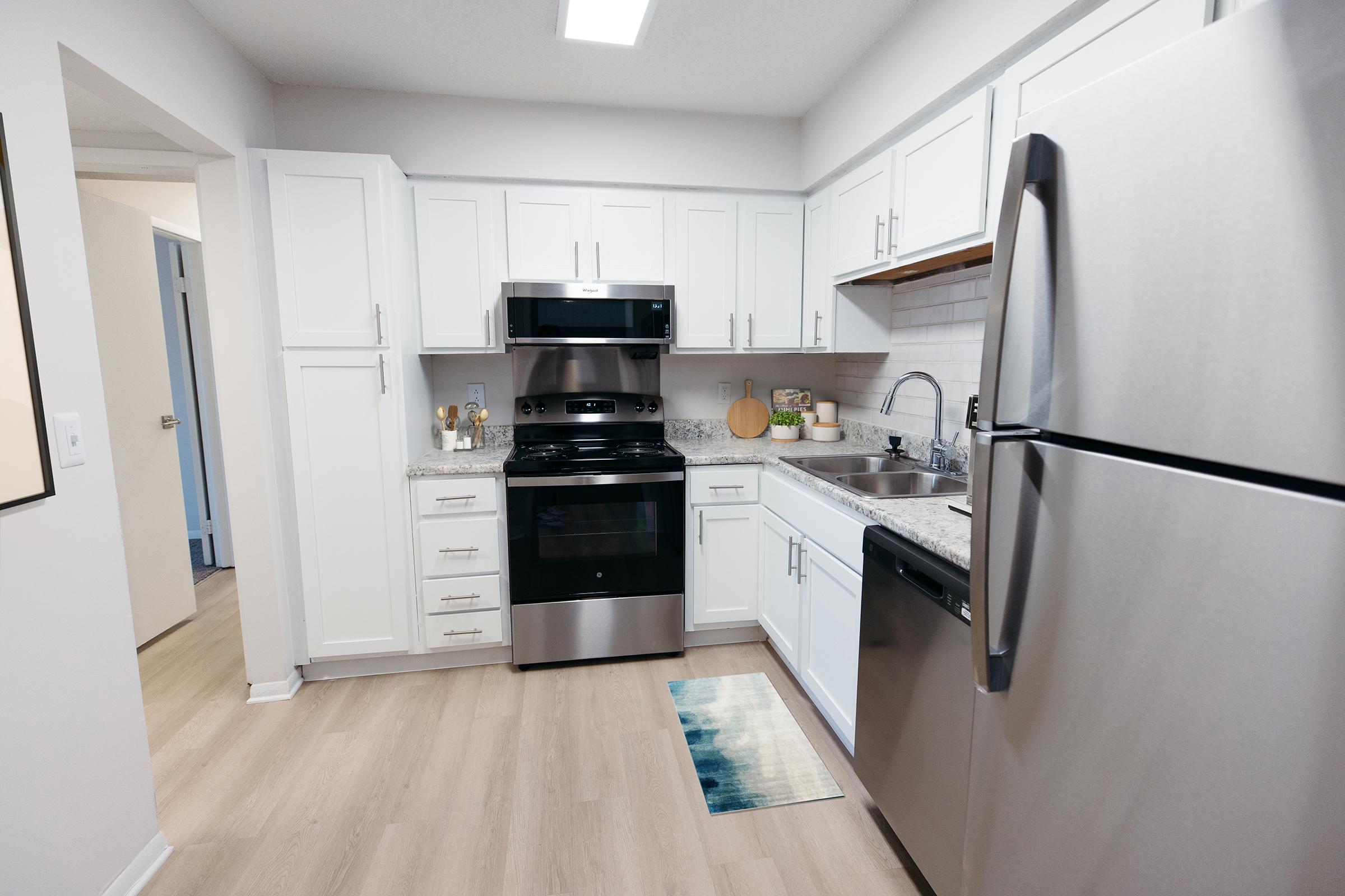 Modern kitchen featuring white cabinetry, stainless steel appliances, and granite countertops. The space includes a stove, oven, sink, and dishwasher, with a wooden cutting board and toiletries on the counter. Light wood flooring and a colorful rug add warmth to the room.
