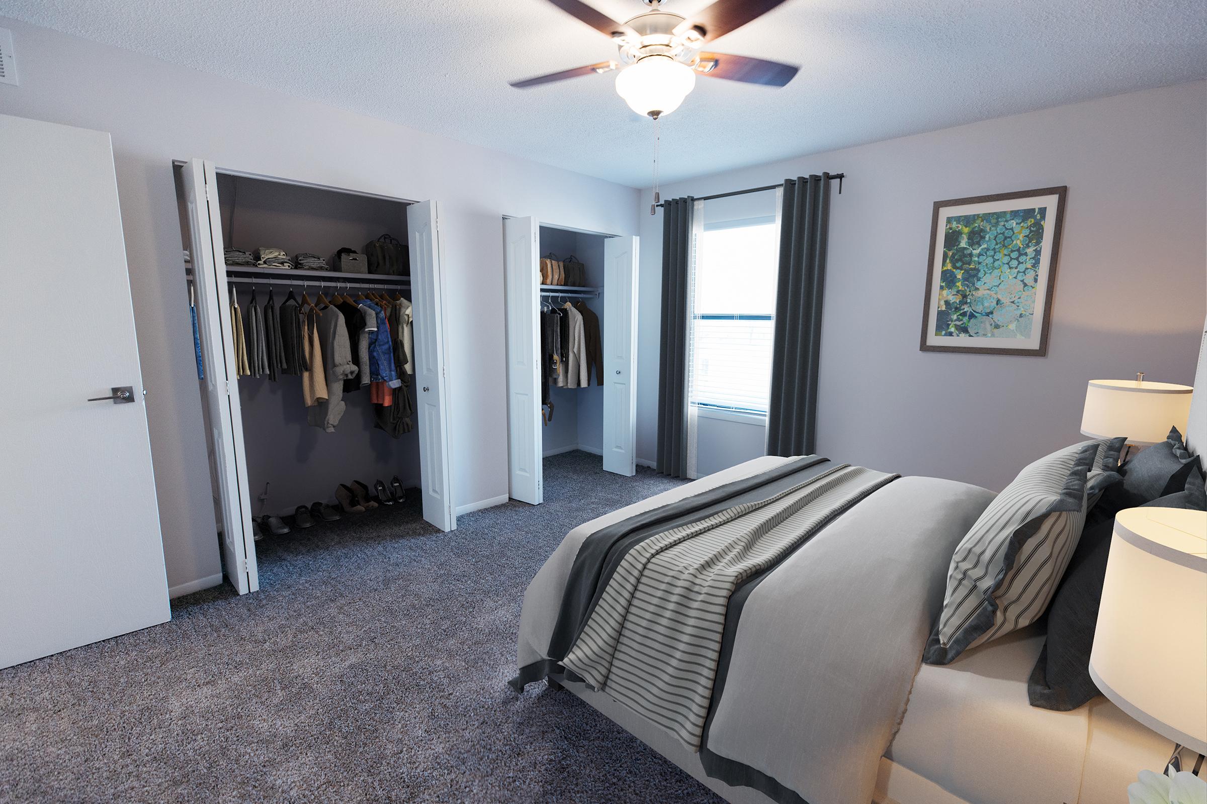 A cozy bedroom featuring a bed with gray bedding and decorative pillows. The room has light purple walls, a ceiling fan, and two closets with various clothing items. A window lets in natural light, and artwork hangs on the wall above the bed. The floor is covered in plush carpeting.