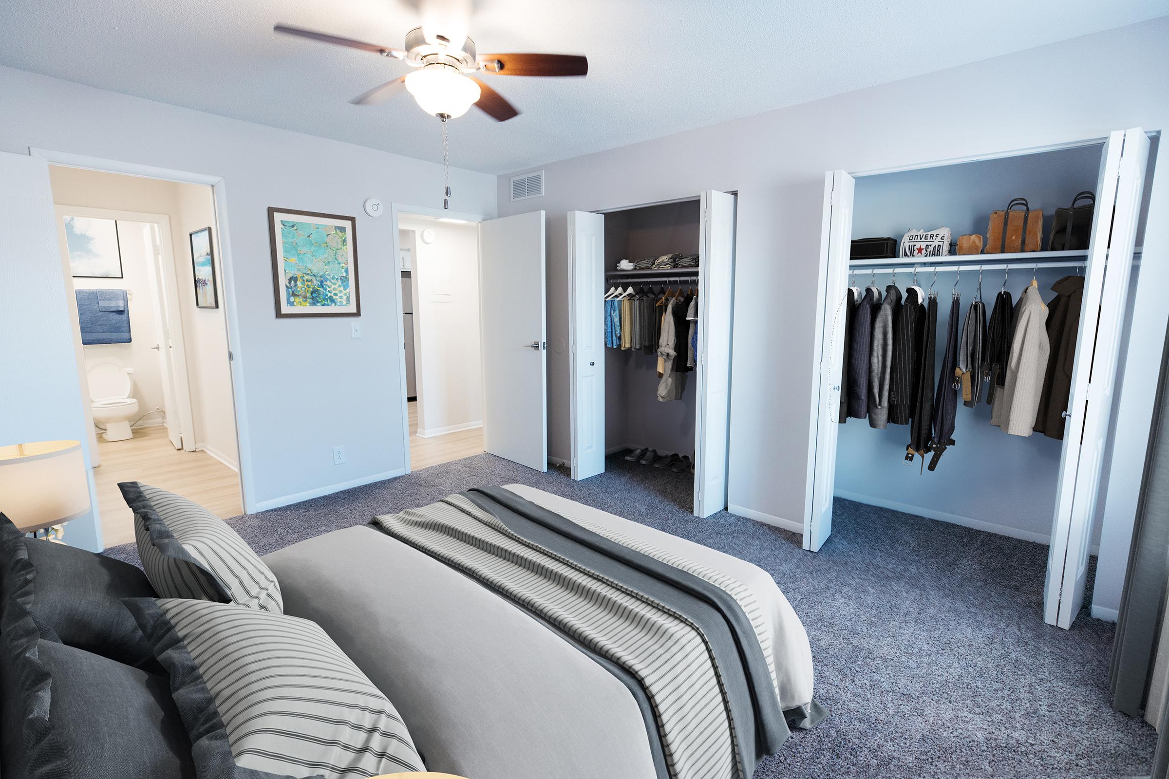 A cozy bedroom with a ceiling fan and a neatly made bed featuring gray and striped bedding. To the sides, there are two open closet doors; one displays a row of hanging jackets, while the other holds bags and accessories. A small bathroom can be seen through a door in the background. The walls are painted light gray.