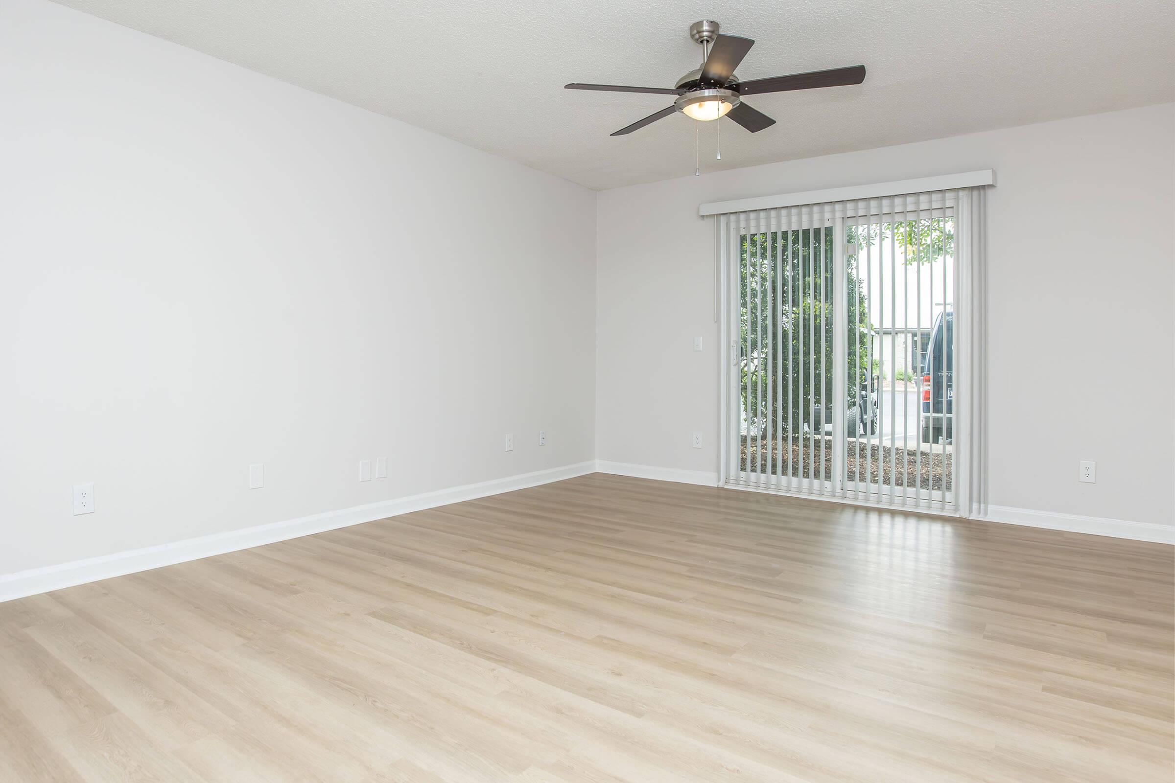 Spacious, empty living room featuring light wood flooring, a ceiling fan, and a sliding glass door with vertical blinds leading to an outdoor area with greenery. The walls are painted in a neutral color, creating a bright and airy atmosphere.