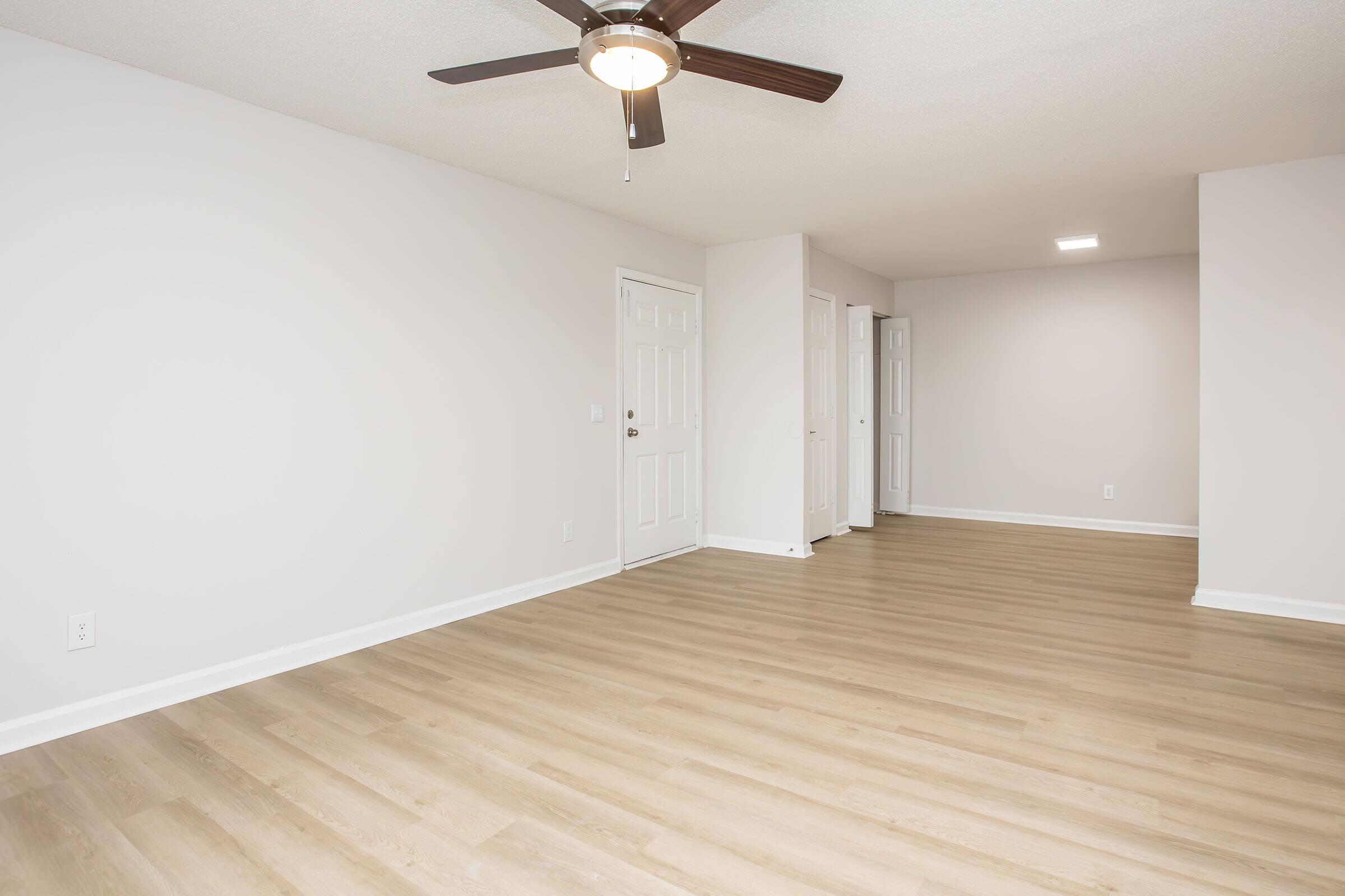 A spacious, empty room with light-colored walls and a light wood floor. There is a ceiling fan with wooden blades, and two closed doors visible on the right side. The room is well-lit, with a soft glow from an overhead light. Ideal for various interior design possibilities.