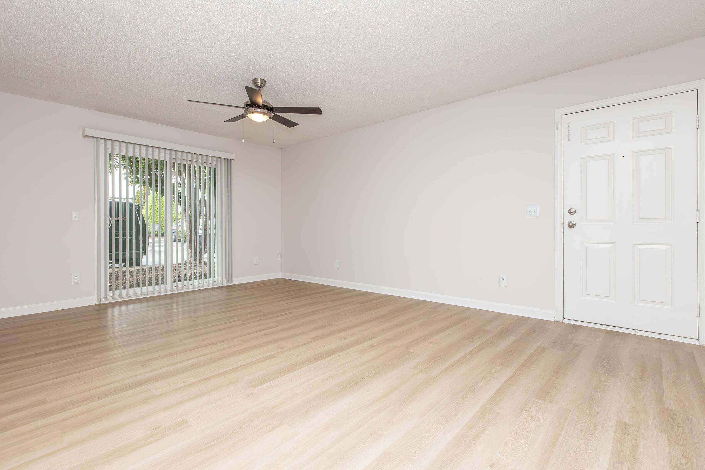 Spacious, empty living room with light-colored walls and wooden flooring. Features a ceiling fan and a sliding glass door with vertical blinds leading to an outdoor area. The front door is on the right side, creating a welcoming entrance. Natural light fills the space, enhancing its openness.