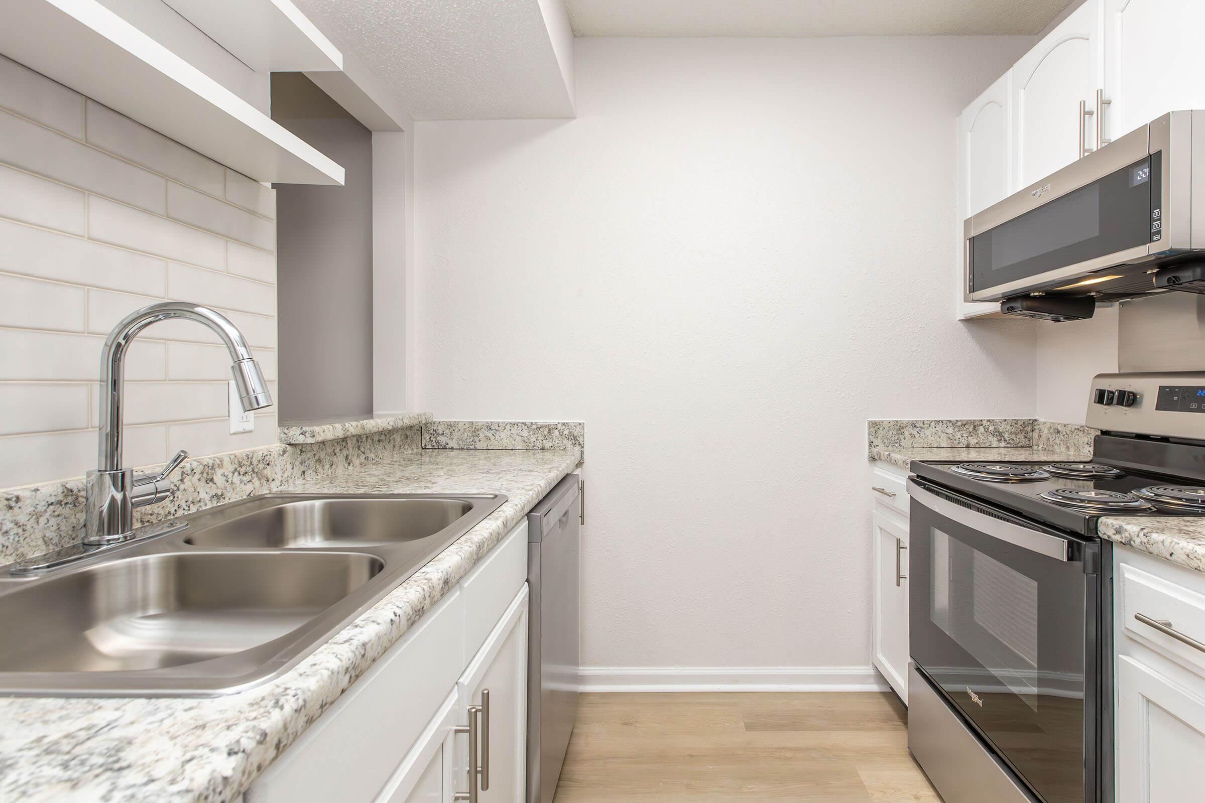 A modern kitchen featuring white cabinetry, a double stainless steel sink, a granite countertop, and stainless steel appliances, including an oven and microwave. The backsplash consists of light-colored tiles, and the flooring is a light wood finish, creating a bright and contemporary atmosphere.