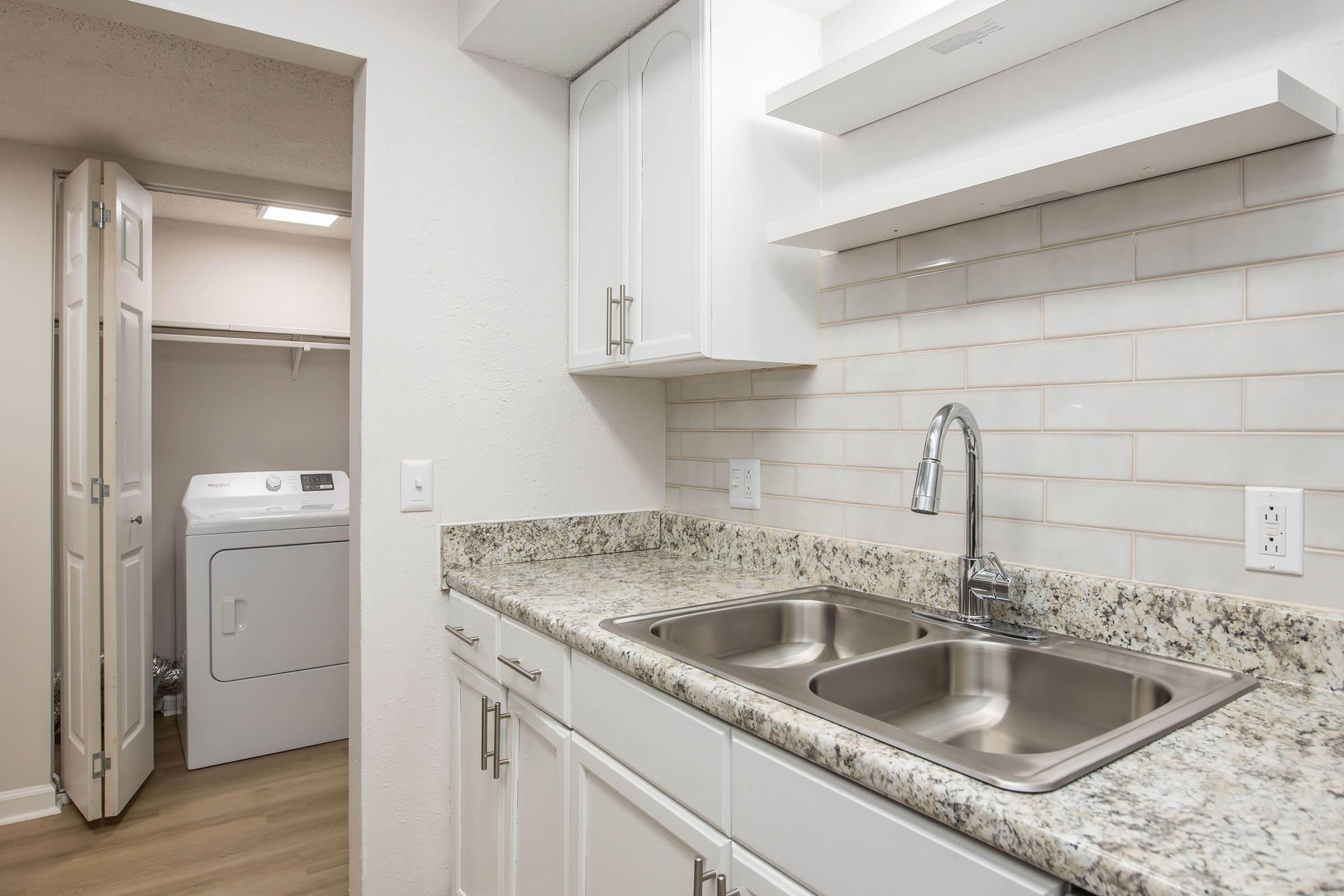 A modern kitchen with a dual sink and granite countertops. White cabinets are installed above and below the sink. In the background, a laundry area is visible with a washing machine and a door leading to another room. The walls are painted in a light color, giving the space a clean and bright feel.