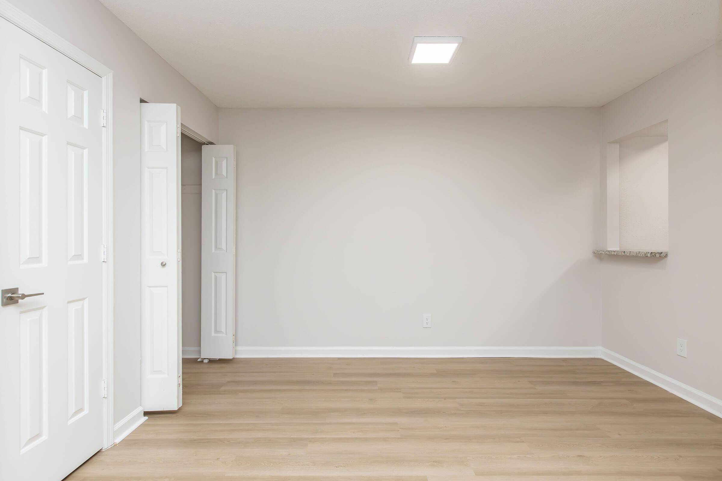 A empty room with light-colored walls and a wooden floor. There are two white doors on the left, one partially open, leading to a closet. A small shelf is built into the wall on the right. The ceiling features overhead lighting, creating a bright, uncluttered space.