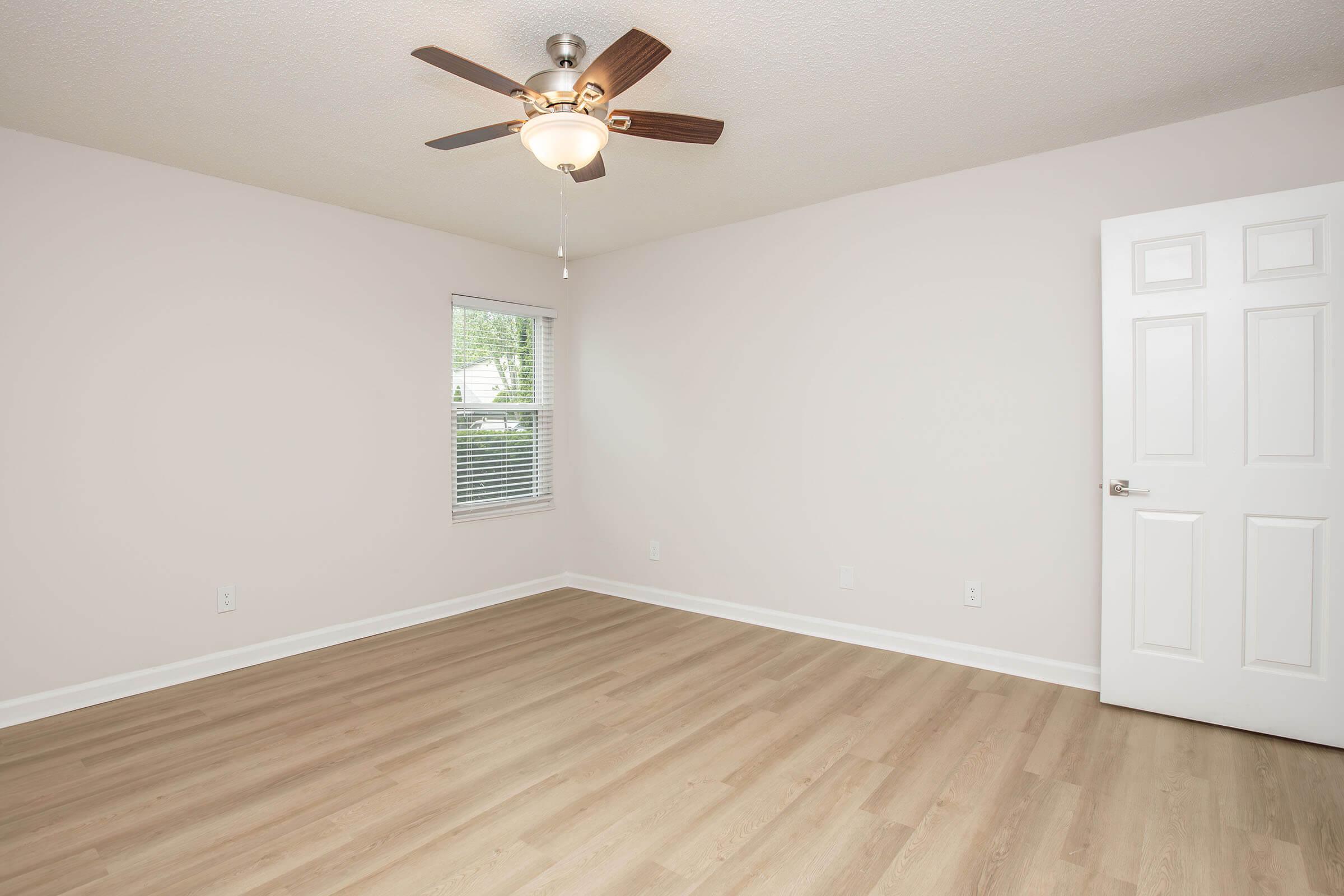 A well-lit, empty room with light-colored walls and a ceiling fan. The flooring is a light wood laminate, and there's a single window allowing natural light. A white door is visible on the right side, leading out of the room.