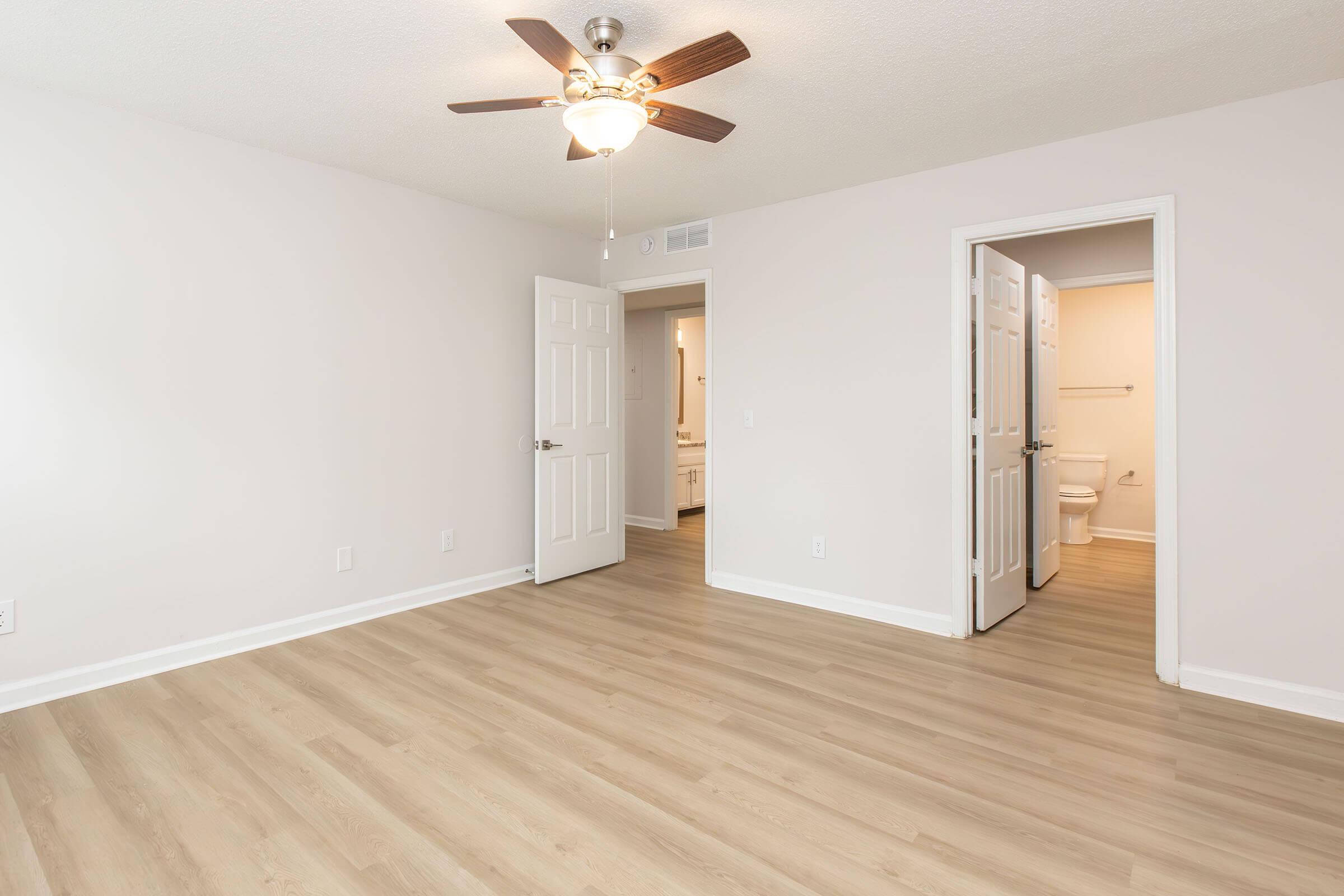 A spacious, empty room with light-colored walls and wooden flooring. A ceiling fan is visible, and there are two open doors leading to bathroom areas. Natural light illuminates the space, creating a bright and airy atmosphere. Ideal for interior decoration or furnishing.