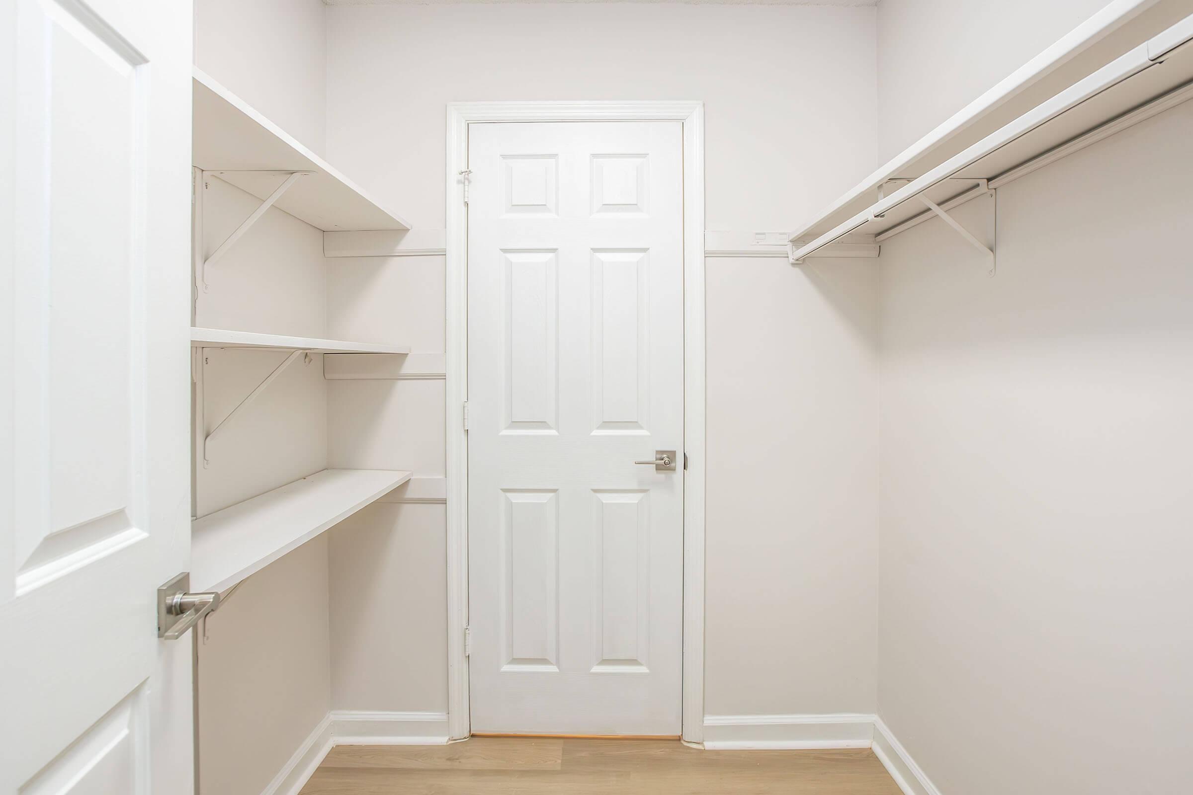A clean and empty closet featuring white walls and natural wood flooring. There are several built-in shelving units on one side and a solid white door leading to another room. The overall design is minimalistic and bright, with plenty of space for storage.