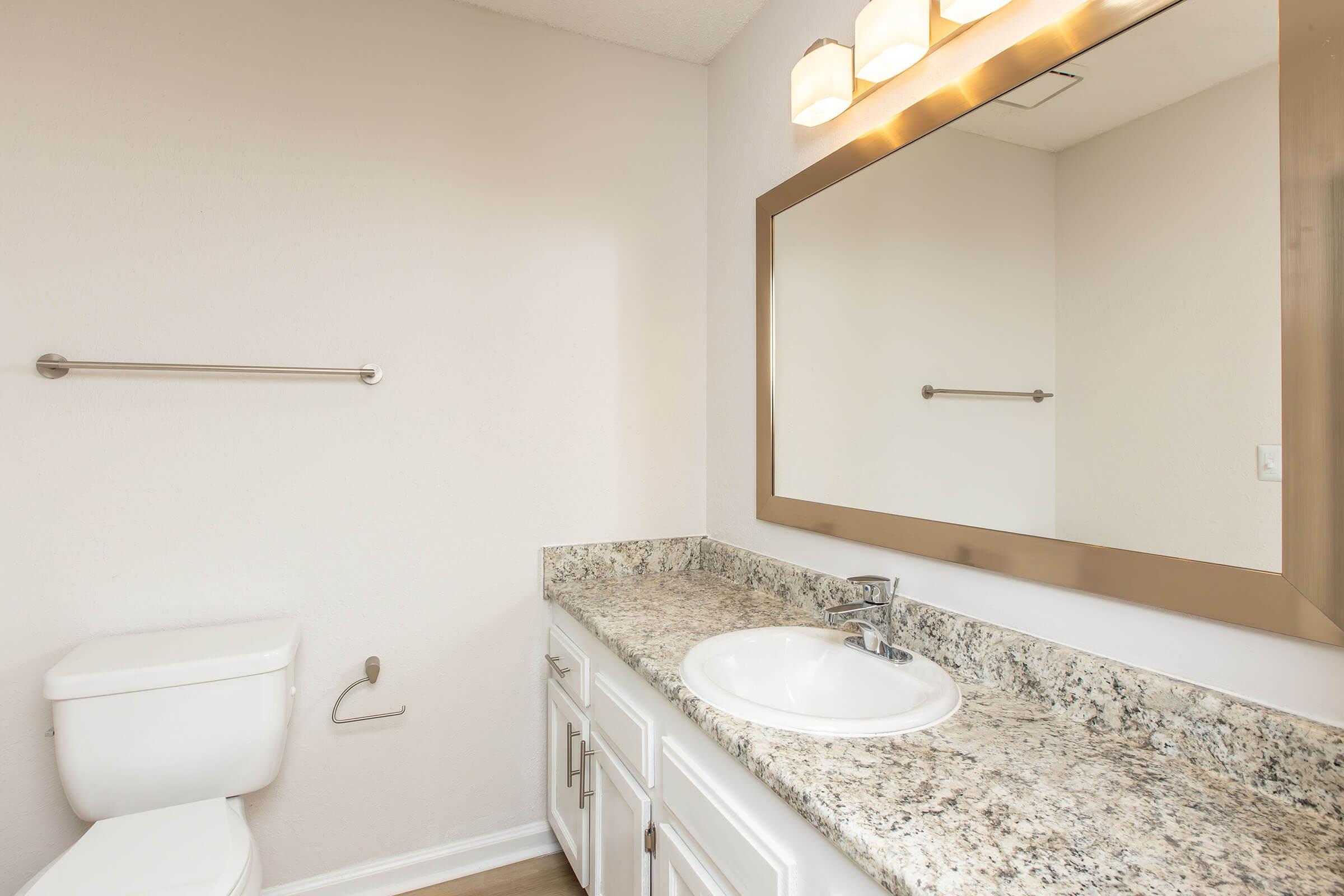 A clean and modern bathroom featuring a white toilet, a granite countertop with a sink, and a large wall mirror. The walls are painted in a light color, and there are three light fixtures above the mirror, providing bright illumination. A towel bar is mounted on the wall beside the sink.