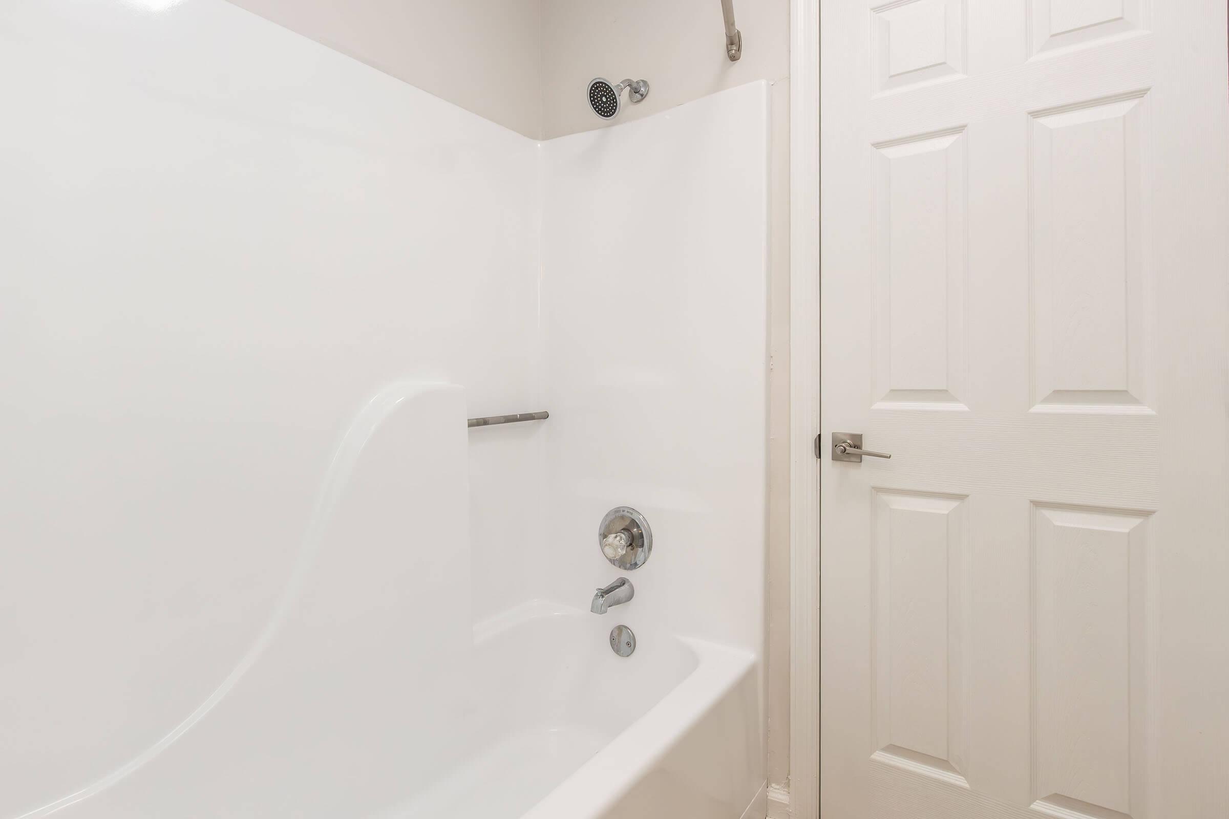 A clean and modern bathroom featuring a white bathtub and wall, a chrome faucet and showerhead, and a closed white door with six panels. The minimalistic design emphasizes light and space, creating a fresh and inviting atmosphere.
