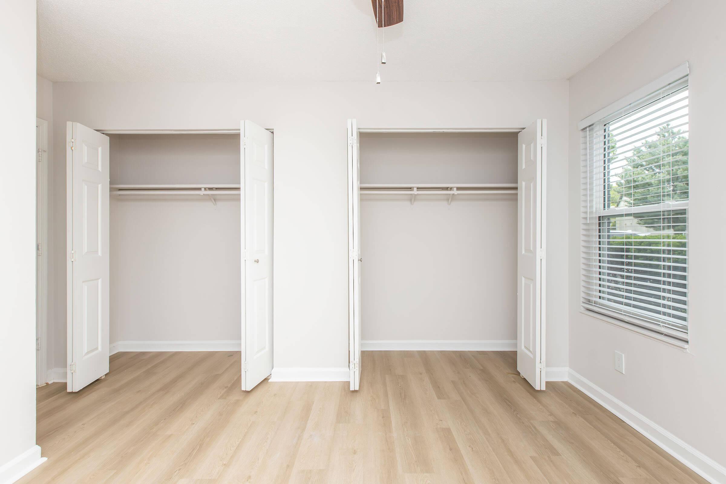 Empty room with two open closets. The walls are painted light-colored, and the floor is light wood. A window on the right features blinds. Natural light enters the space, highlighting its simplicity and cleanliness.