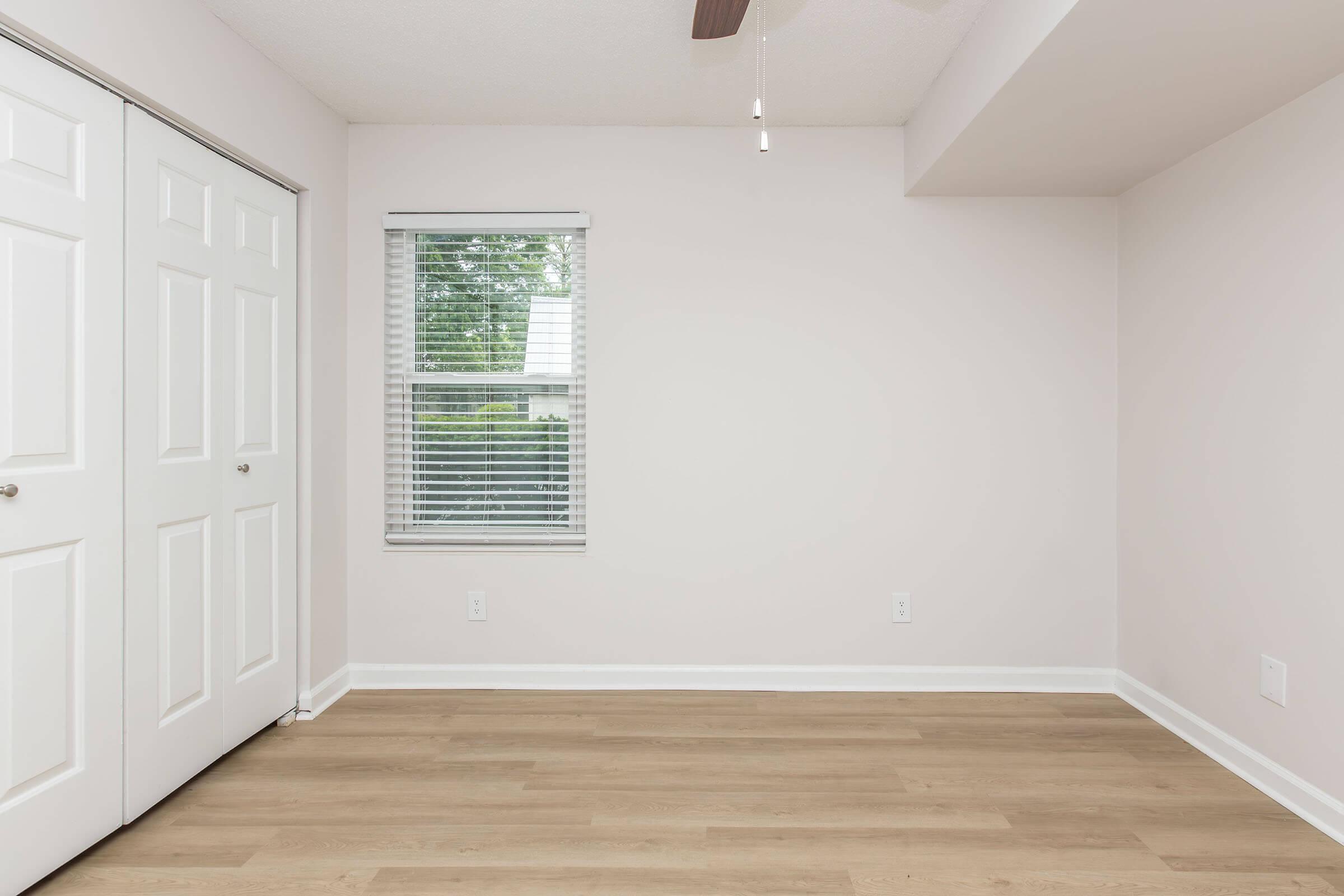 A clean, empty room featuring light-colored walls and wooden flooring. It has a window with blinds on one side and double closet doors on the opposite wall. The ceiling includes a ceiling fan, adding to the bright and airy atmosphere.