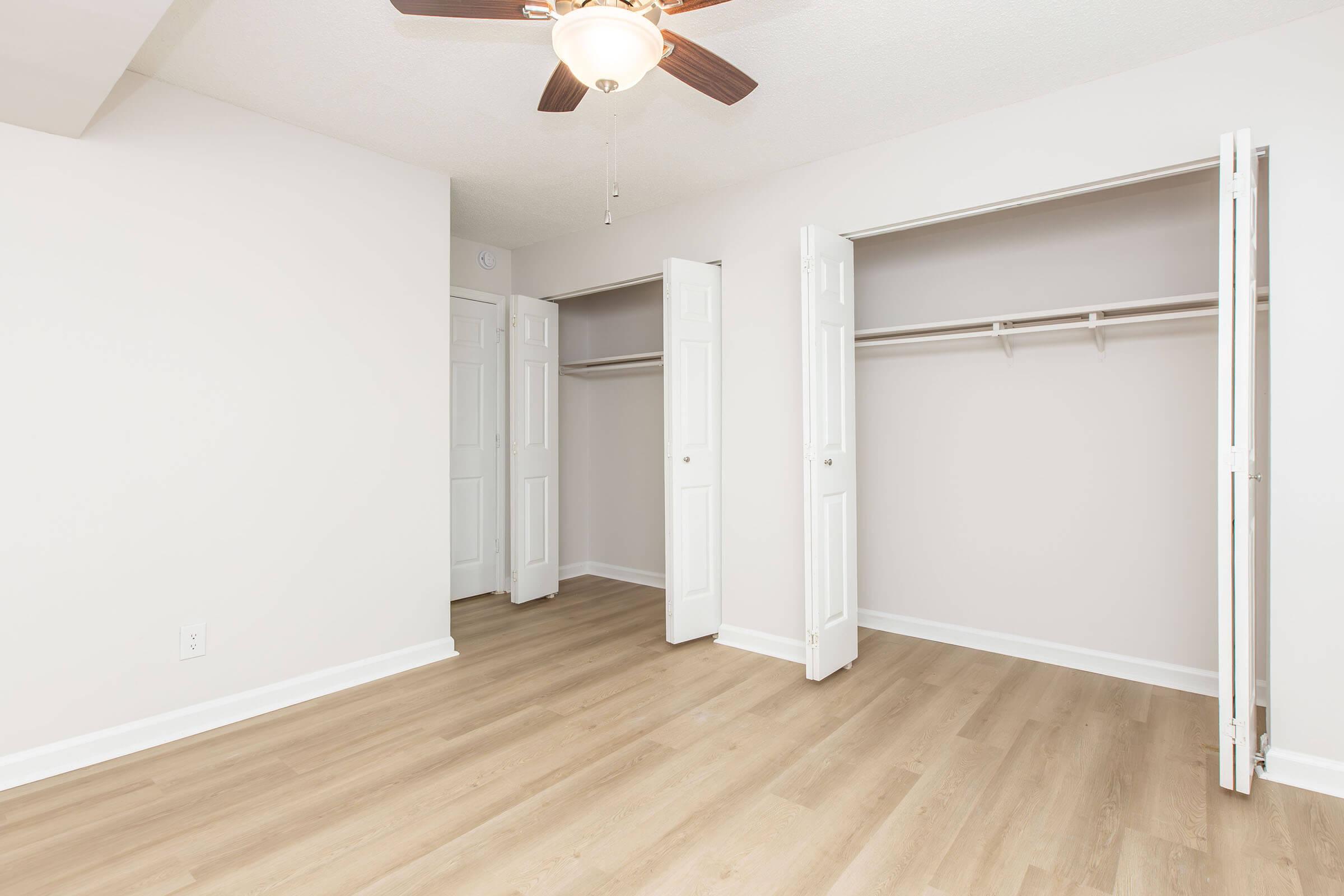 A bright, empty room featuring light-colored walls and hardwood flooring. Two open closet doors on the left reveal hanging space. A ceiling fan is mounted in the center, providing a sense of airiness. The room has a clean and modern feel, making it suitable for various uses, such as a bedroom or office.