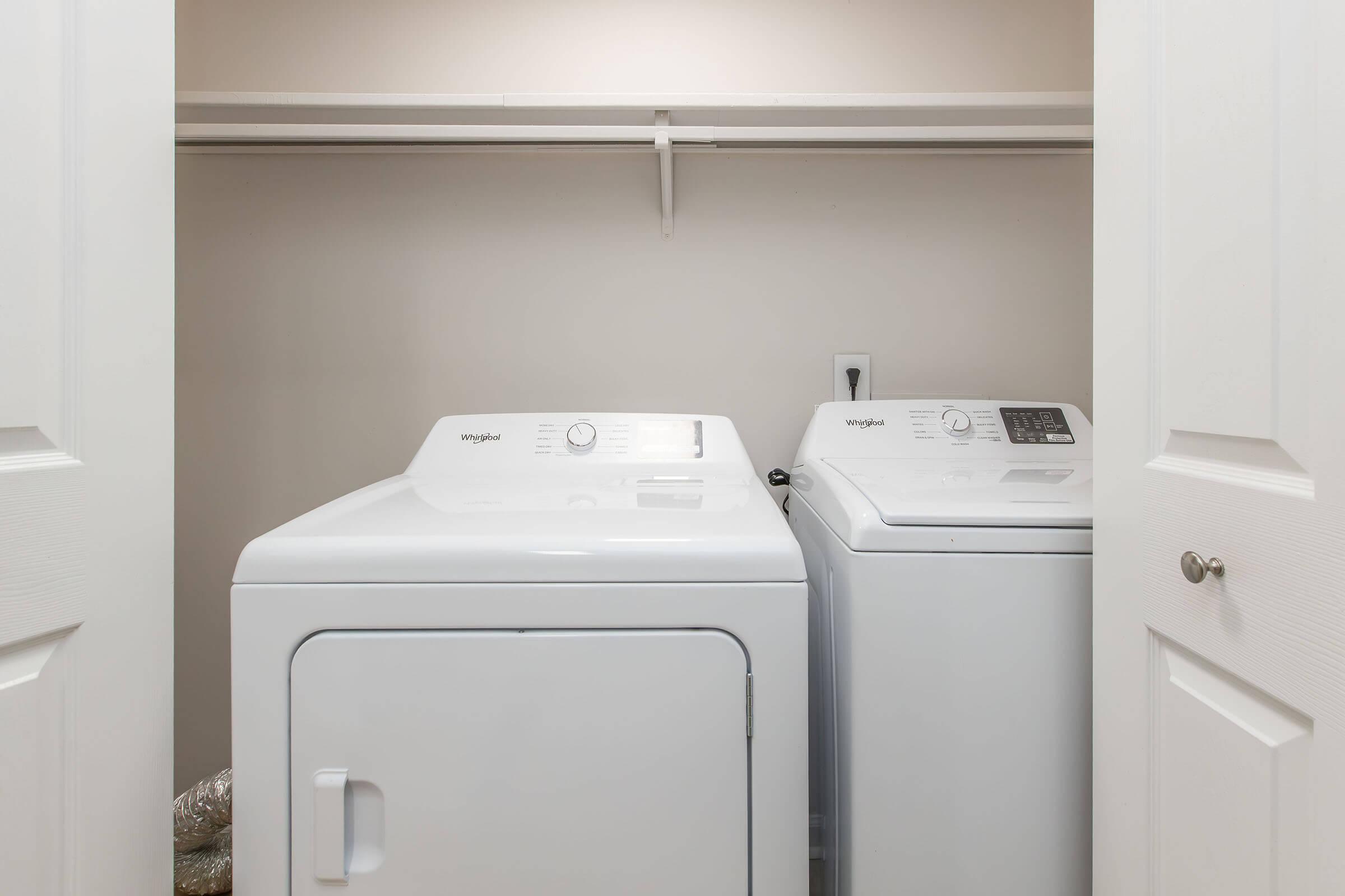 A laundry room featuring a white washing machine and dryer side by side, situated between two closed white doors. The walls are light in color, and there is a shelving unit above the appliances.