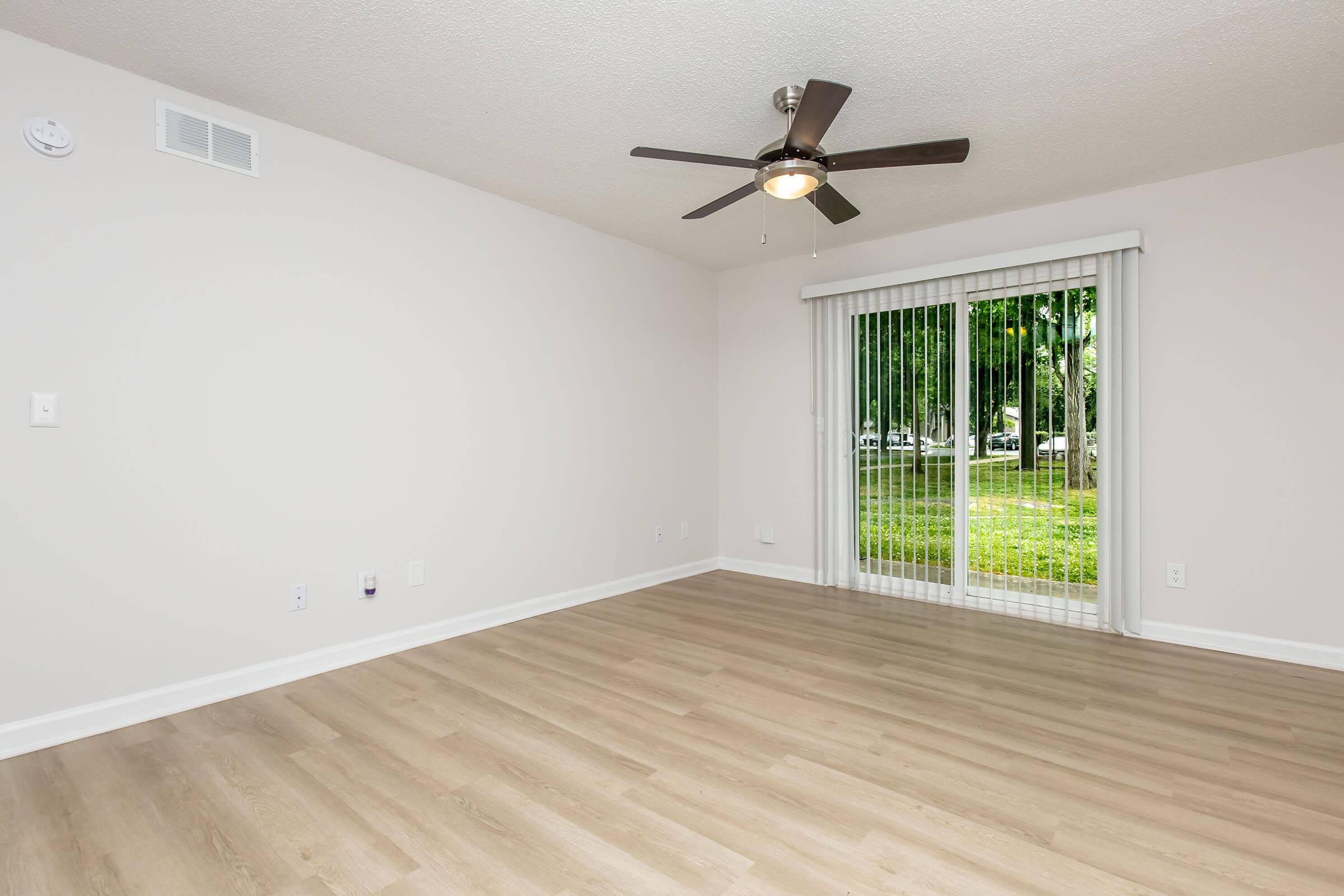 A bright and spacious room with light-colored walls and a light wood floor. There's a ceiling fan in the center, and a large sliding glass door that opens to a view of greenery outside. The room is empty, showcasing a minimalist design with natural light streaming in.