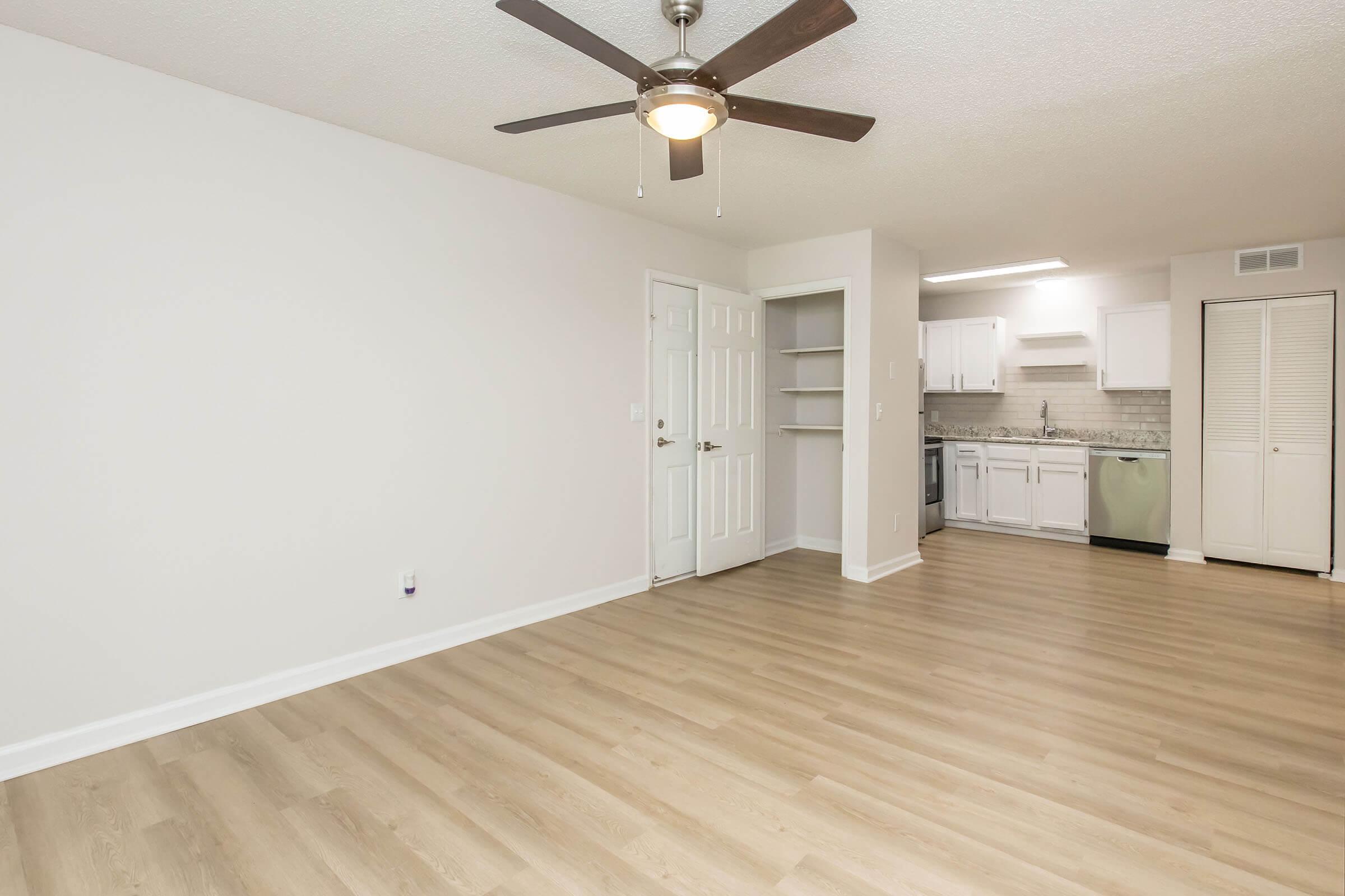 Spacious living area with light hardwood flooring, a ceiling fan, and neutral-colored walls. The open layout features a doorway to a kitchen with white cabinetry, a countertop, and modern appliances. Shelving is visible in the adjacent space, enhancing storage options.