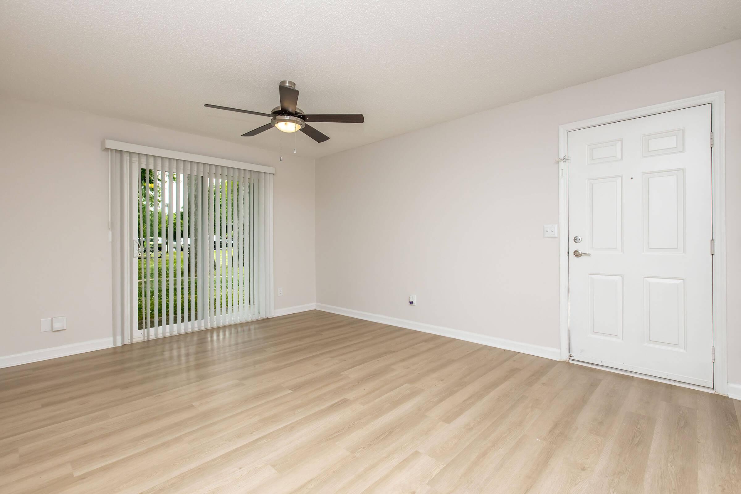 A spacious, empty living room with light wood flooring, a ceiling fan, and a large sliding glass door with vertical blinds that leads outside. The walls are painted a neutral color, and there's a white door leading to another room. Natural light fills the space, creating a bright and welcoming atmosphere.