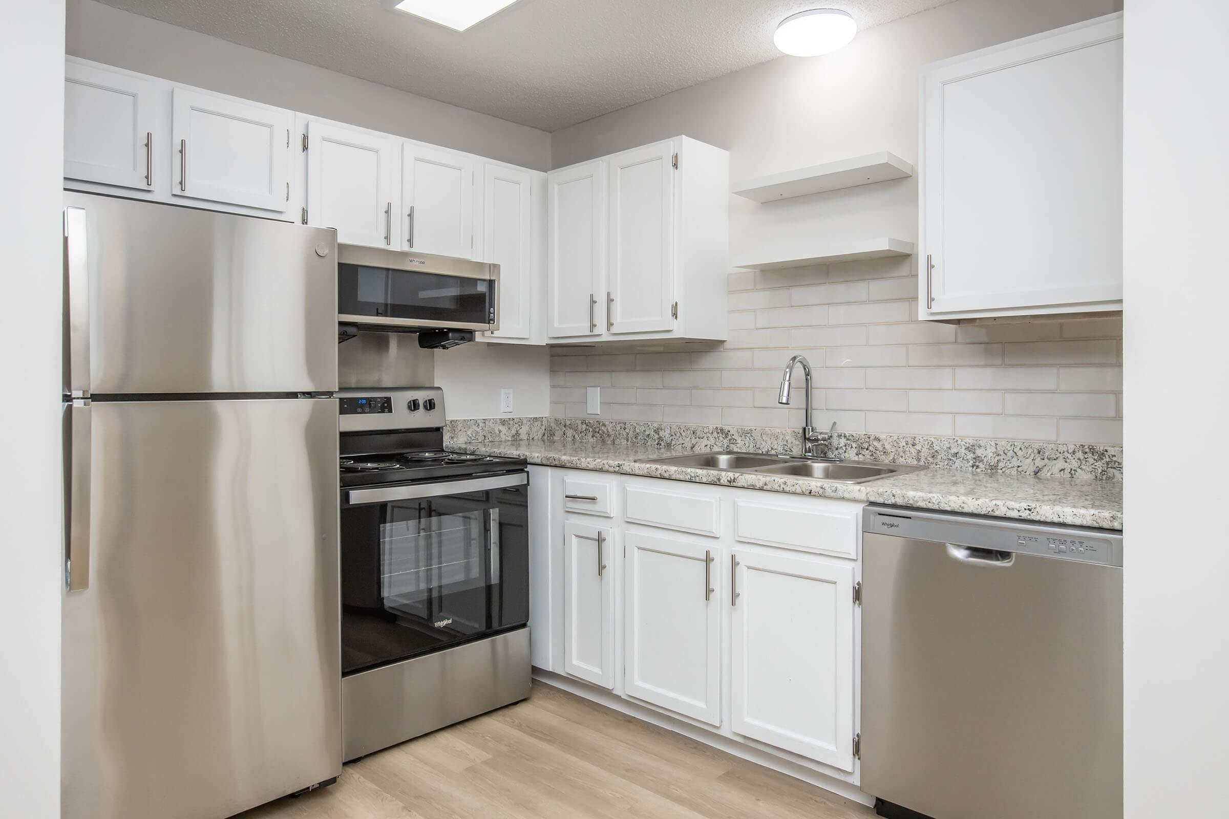 Modern kitchen with stainless steel appliances, including a refrigerator, oven, and dishwasher. It features white cabinetry, a granite countertop, and a light-colored backsplash. The kitchen has a clean, organized layout with a single sink and wood-style flooring.
