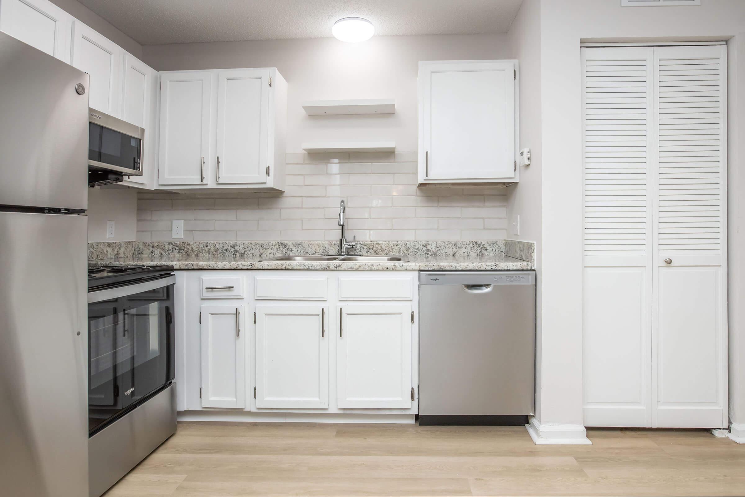 Modern kitchen featuring stainless steel appliances, including a refrigerator, oven, and dishwasher. The cabinets are white with a sleek design, complemented by a light-colored granite countertop. A single sink is installed between the cabinets, with a wall-mounted light fixture above. The floor is a light wood laminate.