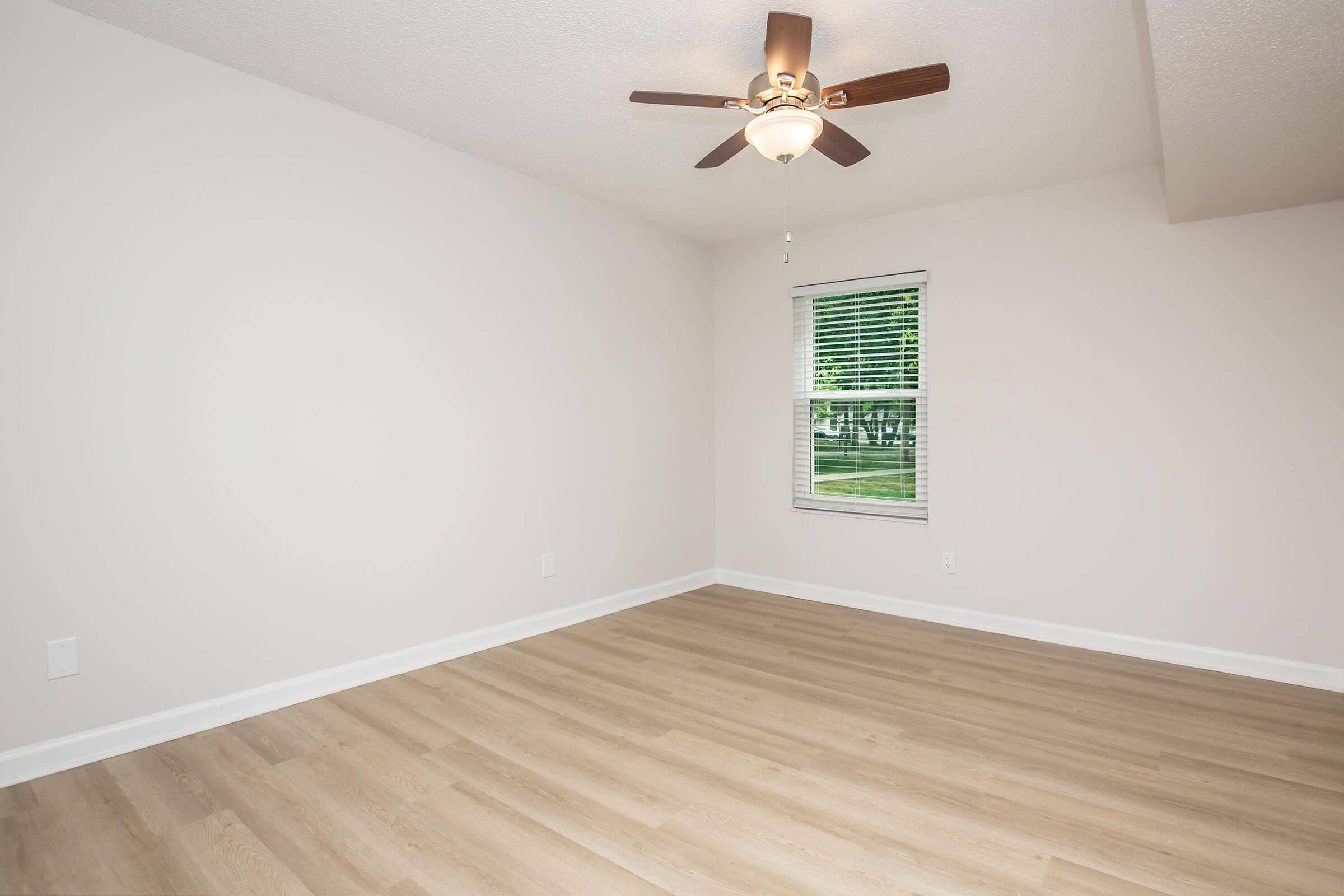 A freshly painted, empty room featuring light-colored walls and a ceiling fan. There is a window with blinds allowing natural light, and the floor is made of light-colored wood laminate. The space is clean and well-lit, ideal for various uses.