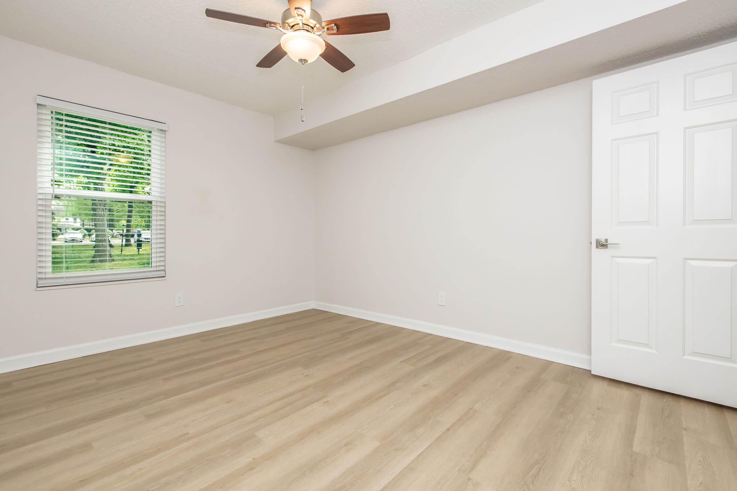 A clean, empty room with light-colored walls and wooden flooring. It features a ceiling fan, a window with blinds allowing natural light, and a closed white door. The room has a spacious feel, making it ideal for various uses.