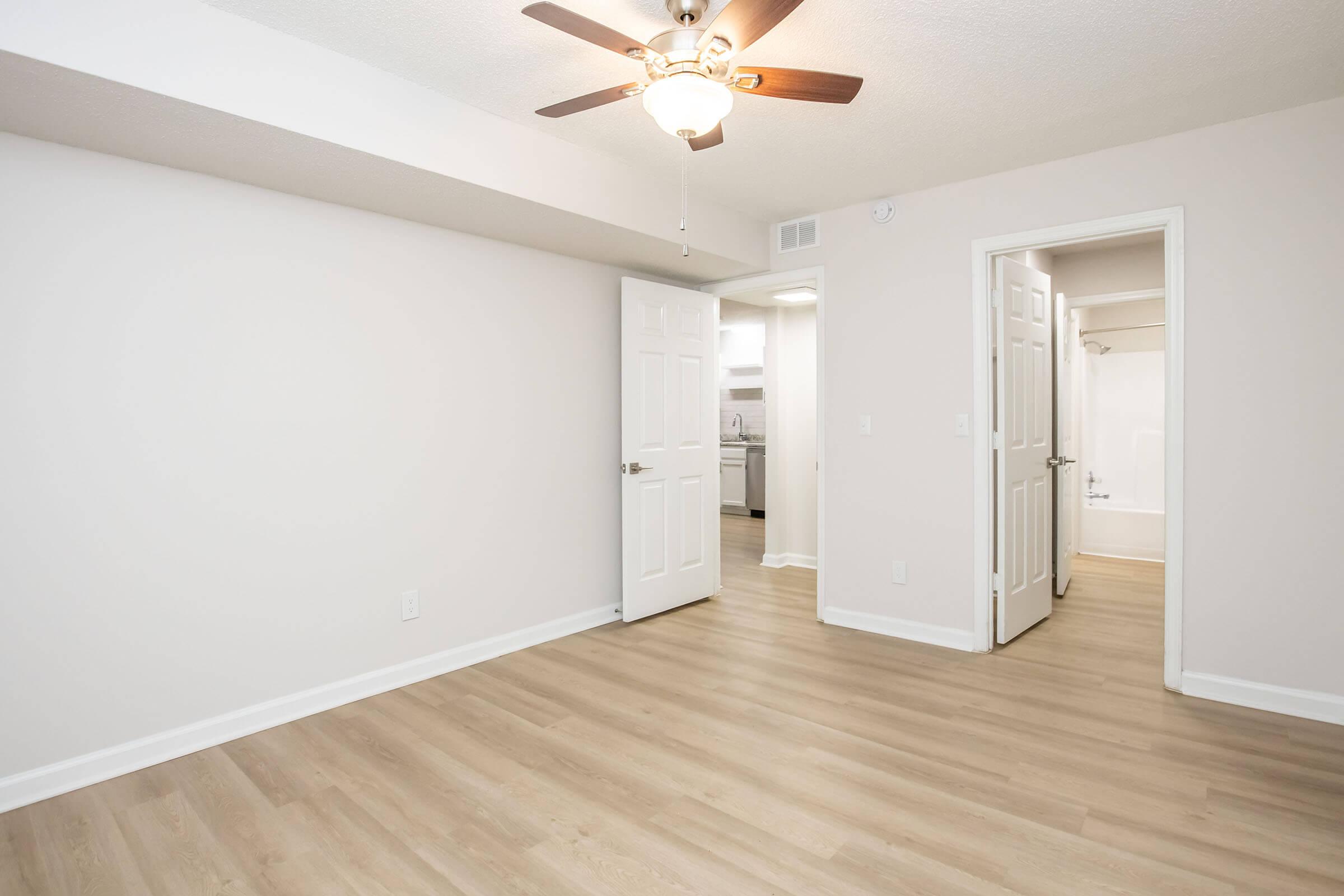 A well-lit room featuring light-colored walls and wood flooring. There is a ceiling fan and an open door leading to a bathroom. A second door is visible, leading to an adjacent area, possibly a kitchen. The space appears clean and modern, suitable for various uses.