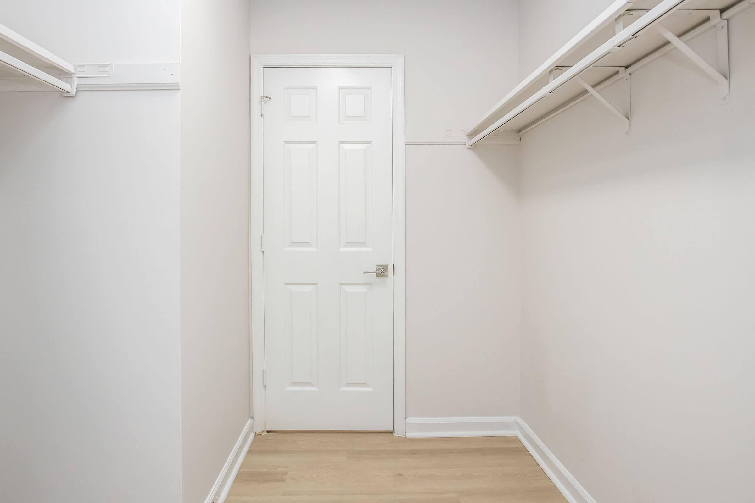 Empty closet space with light-colored walls and wooden flooring. A white door is visible at the end of the closet, and there are two empty shelves along the side walls. The room is well-lit, giving a clean and spacious appearance.