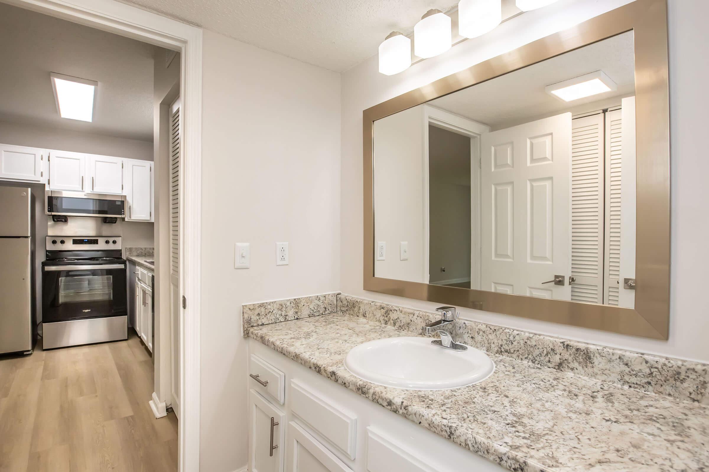 A modern bathroom featuring a granite countertop with a sink, a large mirror above it, and bright lighting. The background shows an open kitchen area with stainless steel appliances and white cabinetry, creating a harmonious and spacious living environment.