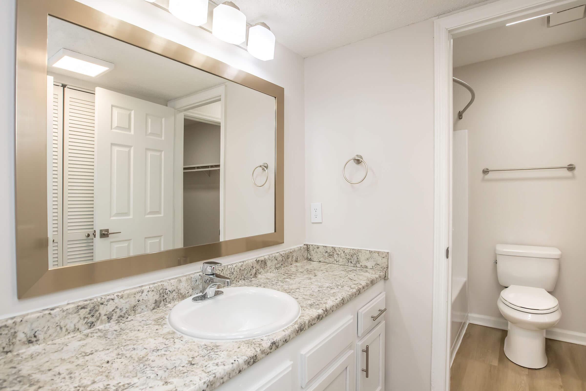 A well-lit bathroom featuring a granite countertop with a sink, a large mirror framed in brushed metal, a towel ring, and a sleek white toilet. The space has light-colored walls, a closet door visible in the background, and a shower area, creating a clean and modern aesthetic.
