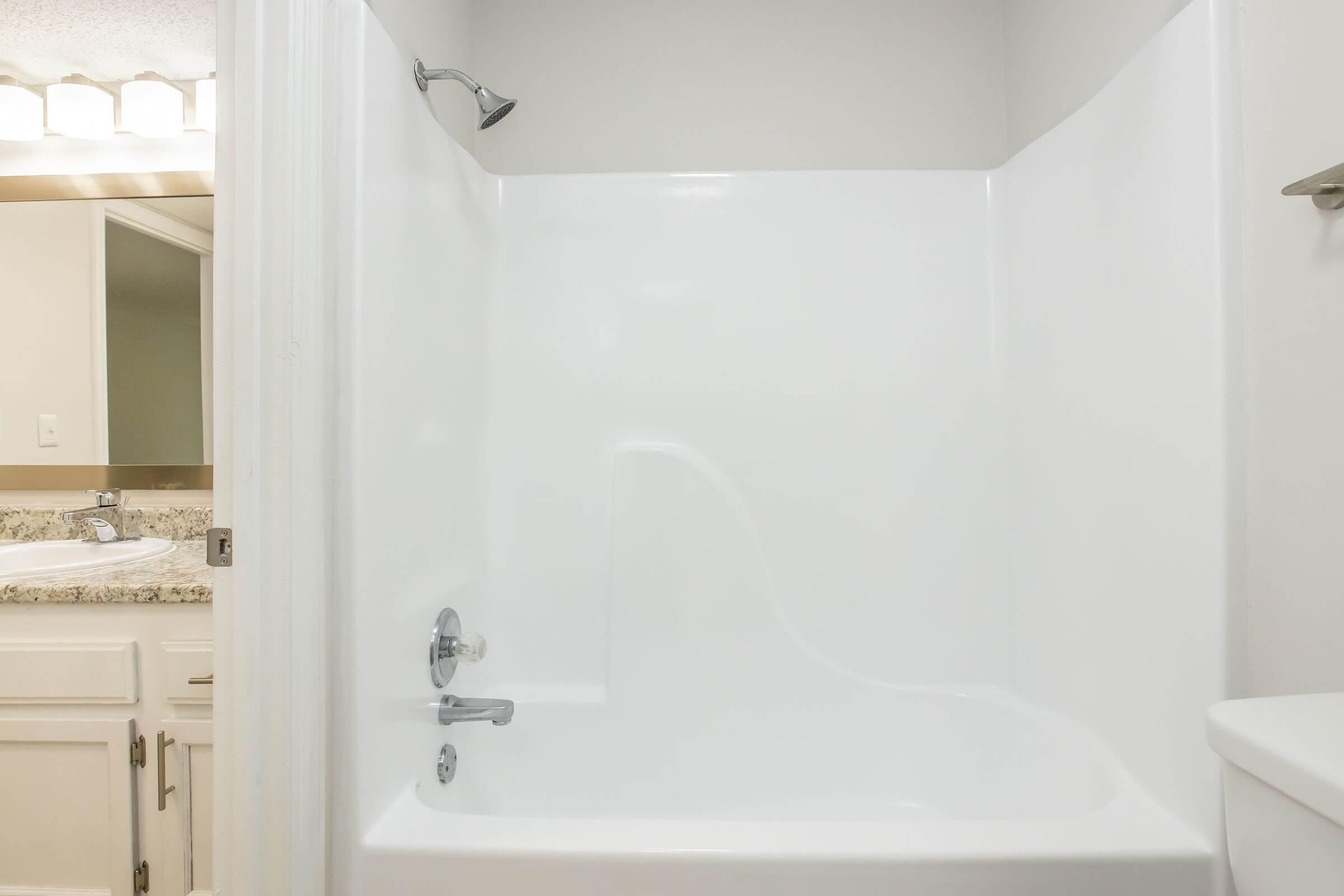 A clean, white bathtub and shower area with a chrome showerhead. The surrounding walls are smooth and feature a minimalist design. A small section of a bathroom sink and mirror is visible in the background. The overall aesthetic is modern and bright.