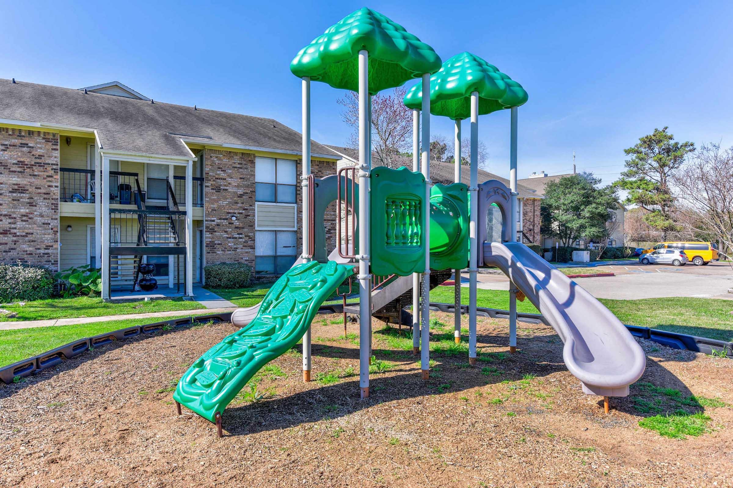A playground featuring two green slides and a structure with a leafy design. It's set on a grassy area with mulch and surrounded by apartment buildings, providing a recreational space for children. Bright blue sky and trees are visible in the background.