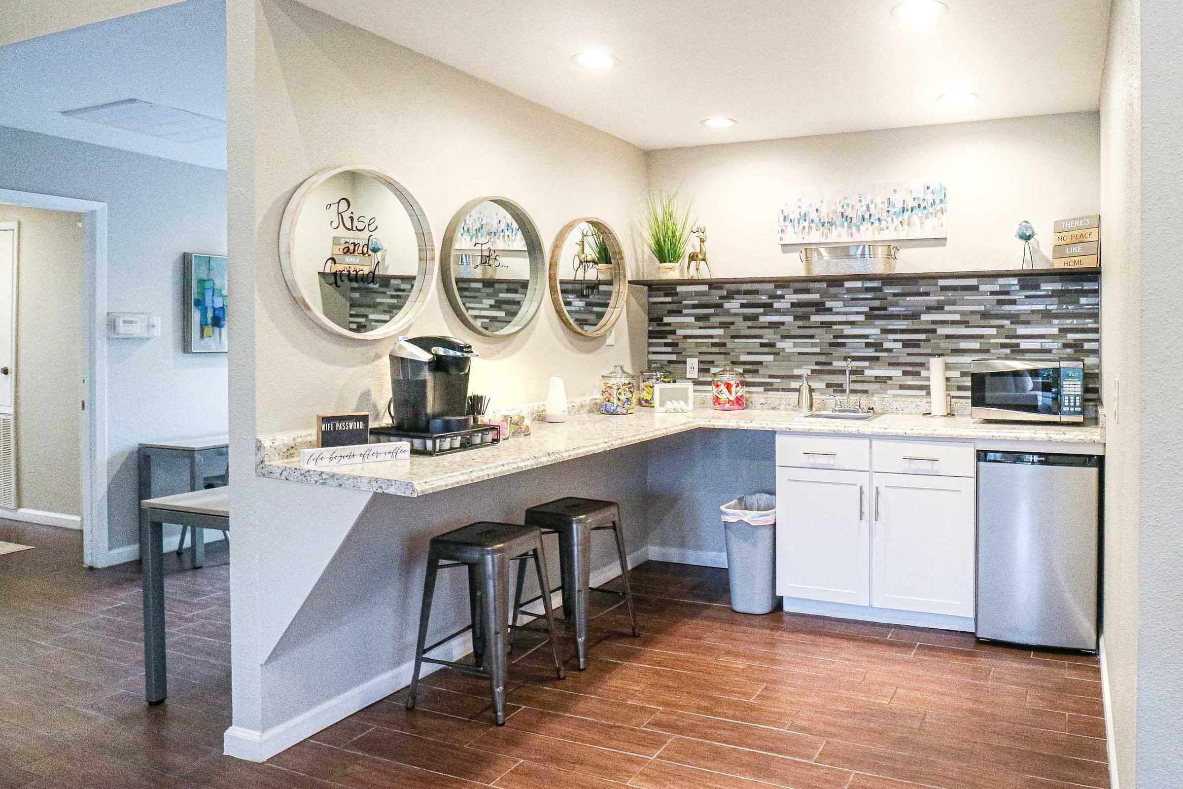 Modern kitchen with a granite countertop, featuring three circular mirrors on the wall, a coffee maker, and a stainless steel sink. The backsplash has a mosaic design, and there are bar stools at the counter. A small trash can is visible, along with decorative items and plants on the shelves.