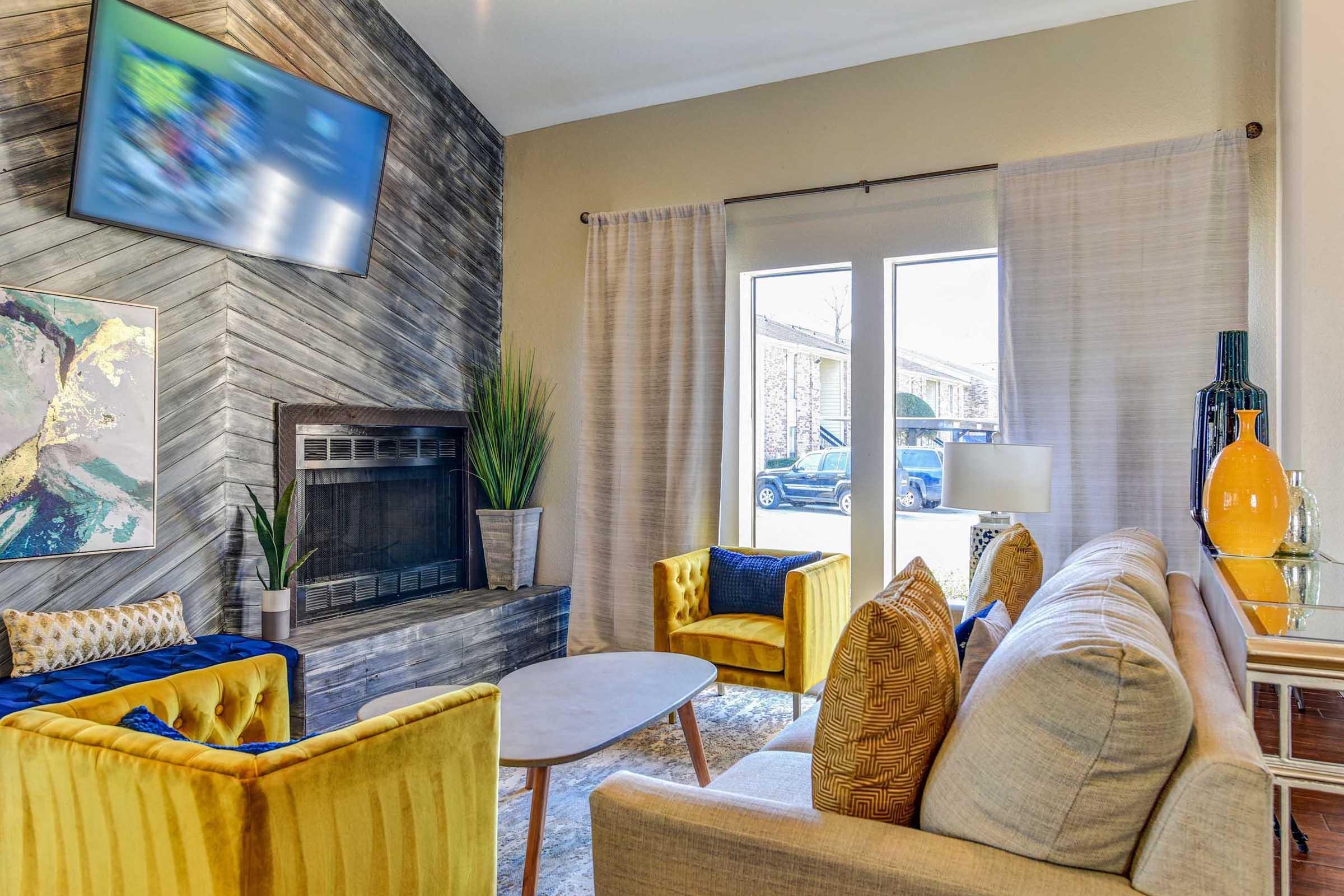 A cozy living room featuring a modern design. It has a light-colored sofa with decorative pillows, two yellow accent chairs, a coffee table, and a fireplace with a wooden accent wall. Large windows allow natural light, revealing a view of a parked car outside. Two curtains frame the windows. A flat-screen TV is mounted on the wall.