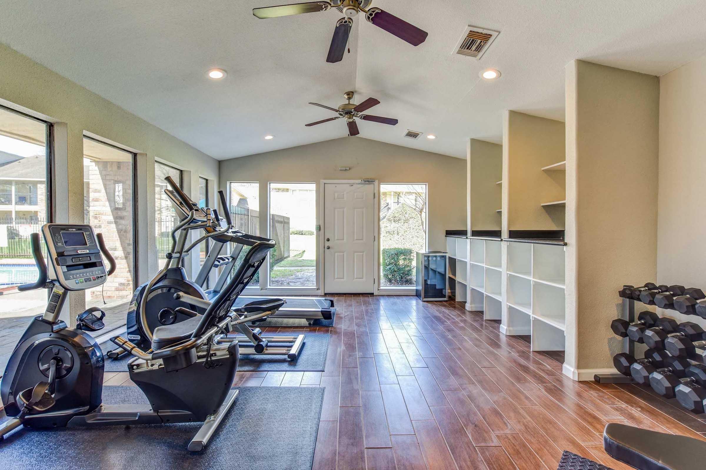A well-lit gym area featuring exercise equipment including a stationary bike and elliptical machine, with a wall of weights. Large windows provide natural light, and there is a view of an outdoor area. The room has wooden flooring and contemporary decor.