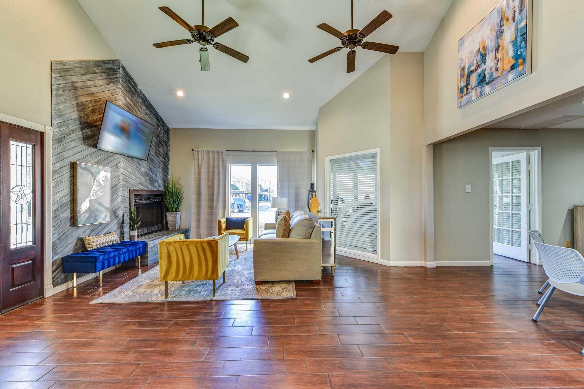A spacious living room featuring a high ceiling, wooden flooring, and large windows. There are two ceiling fans, a blue sofa, a yellow armchair, and a beige sofa arrangement. A flat-screen TV is mounted on the wall above a fireplace. Decor includes a modern artwork piece and decorative plants.