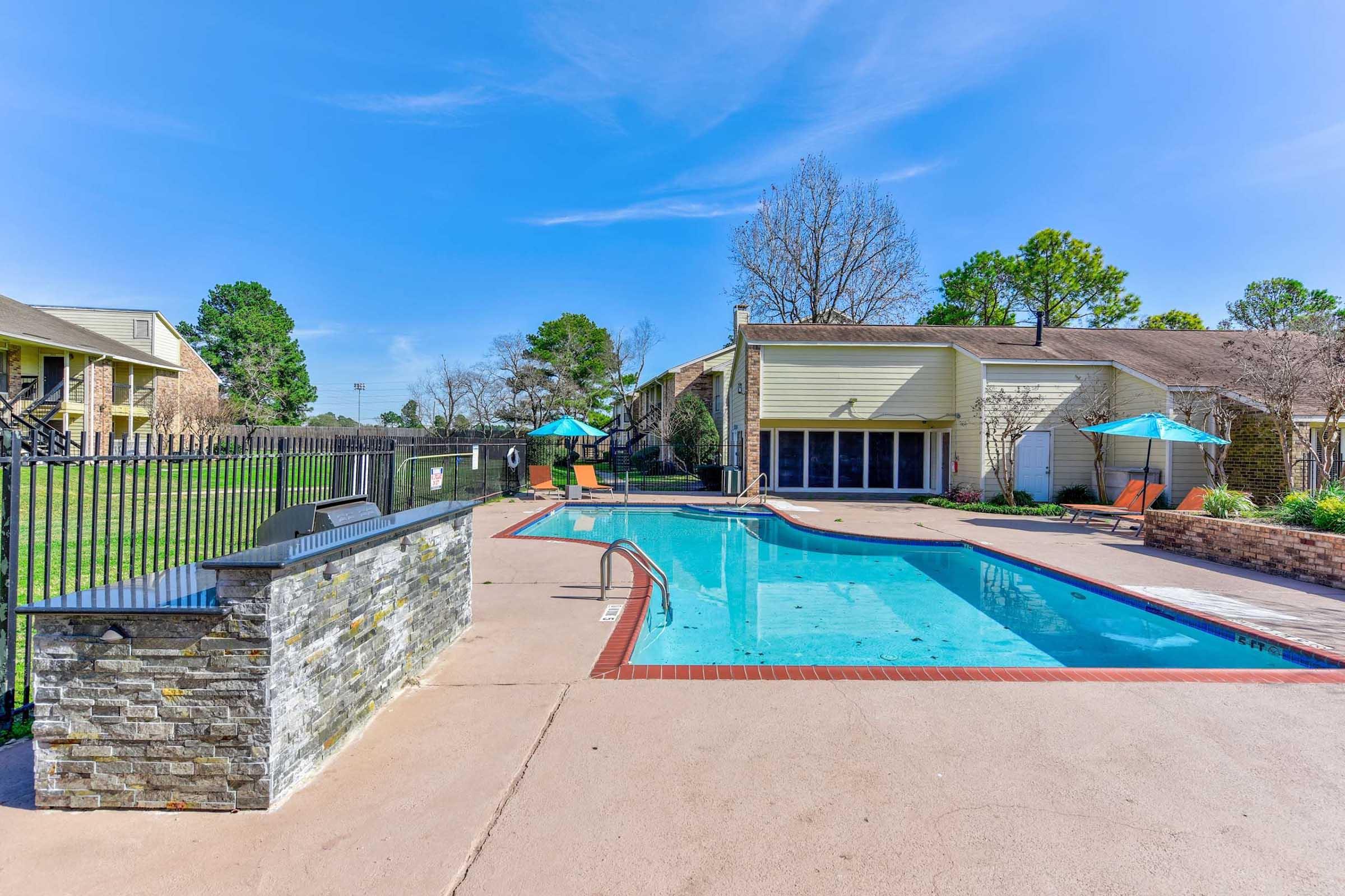 A clear blue swimming pool surrounded by a concrete patio, with lounge chairs and umbrellas. There is a stone wall near the pool, and the area is enclosed by a fence. In the background, grass and trees are visible, along with a building. The sky is bright blue with a few clouds.