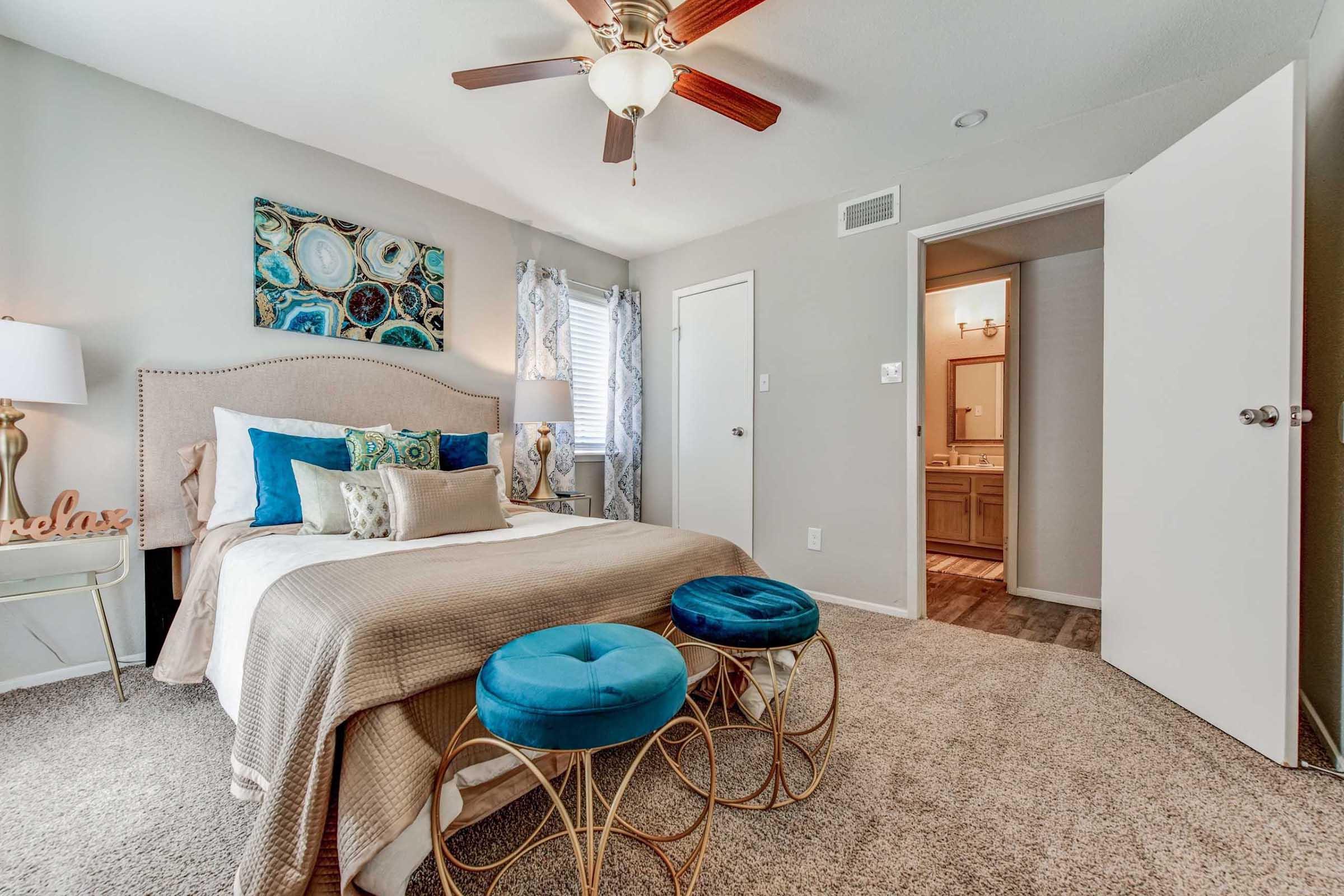 A modern bedroom featuring a plush bed with decorative pillows, two round blue ottomans, and a stylish lamp. The walls are adorned with abstract artwork, and there's a doorway leading to a bathroom area. Soft lighting and neutral colors create a cozy atmosphere.