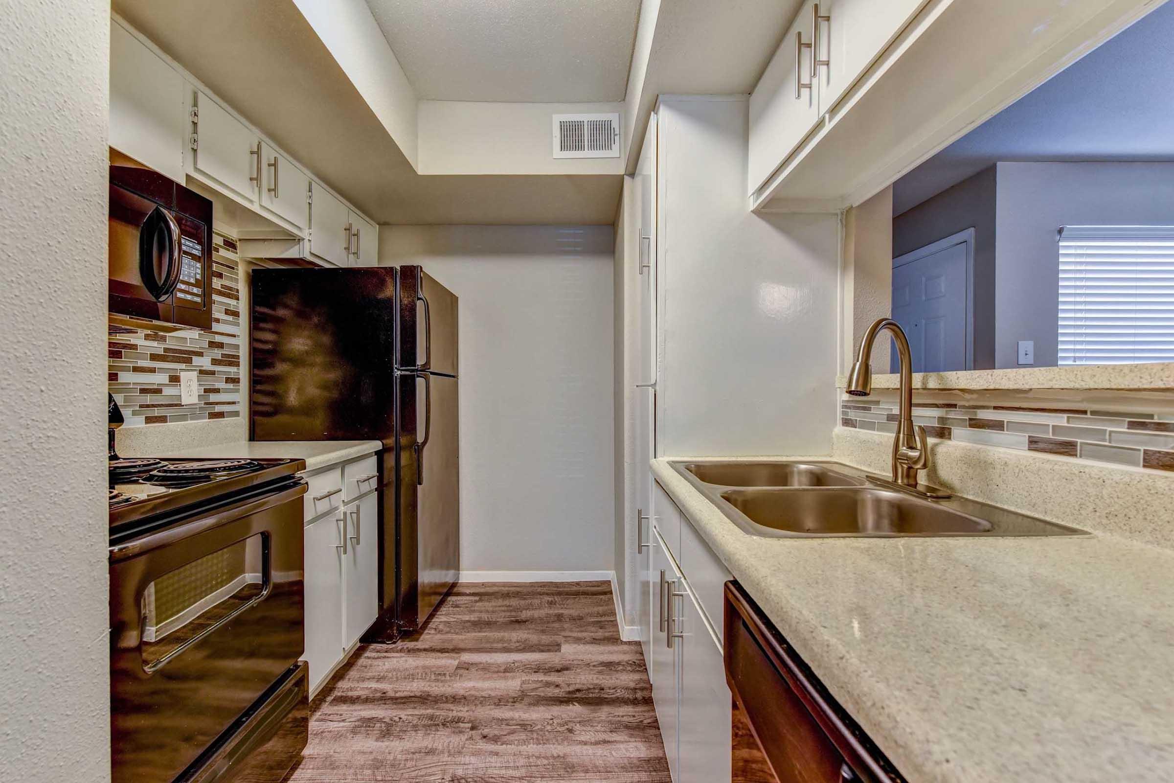 Modern kitchen with dark appliances, including a refrigerator and stove. Light-colored cabinets with a textured backsplash complement the granite countertop. A double sink is located next to the microwave and stove. The space features wood-style flooring and a window providing natural light.