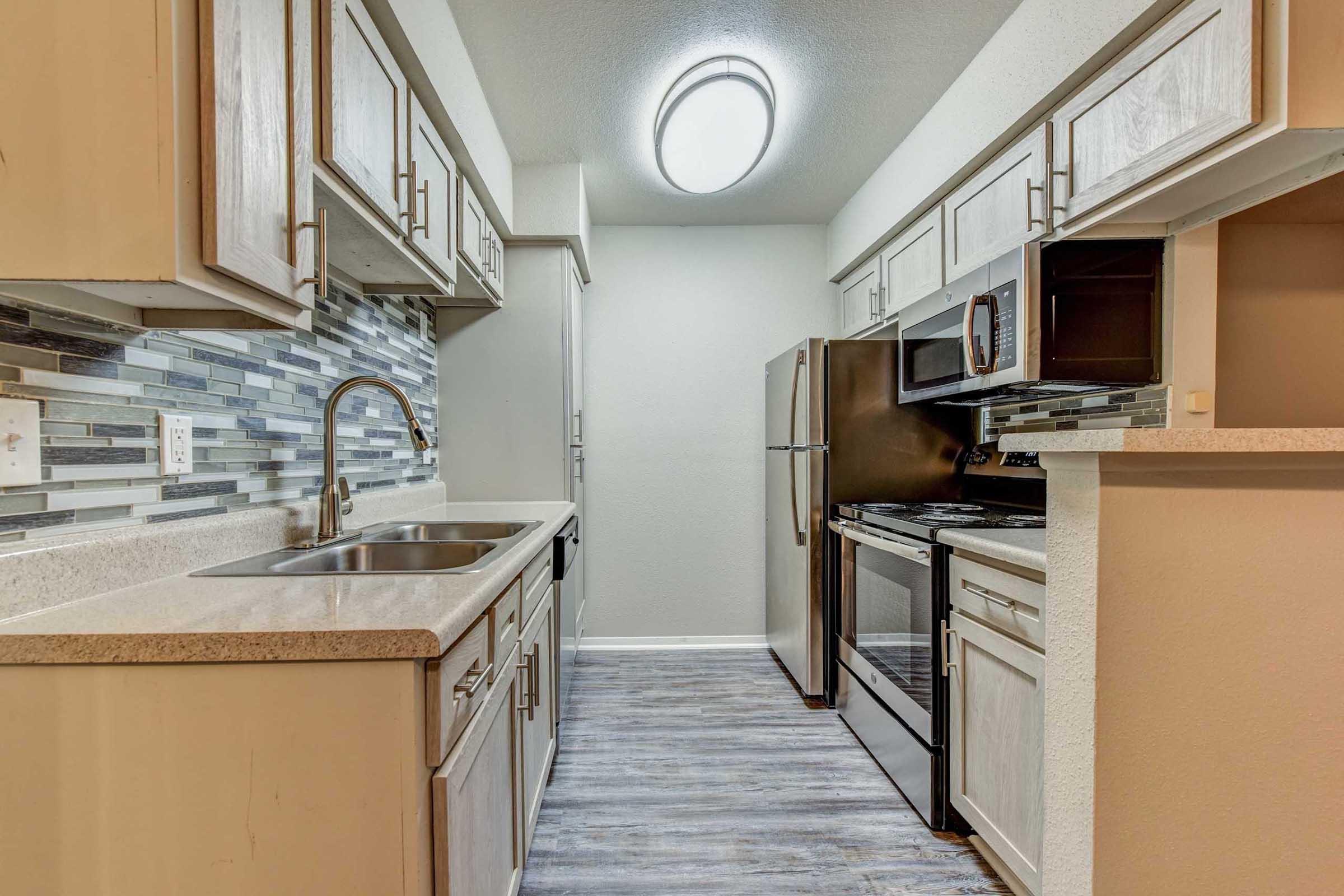 Modern kitchen with light-colored cabinets, stainless steel appliances, and a sleek countertop. Features a sink, stove, and microwave, with a decorative tile backsplash. The space is well-lit with overhead lighting, creating a bright and inviting atmosphere.