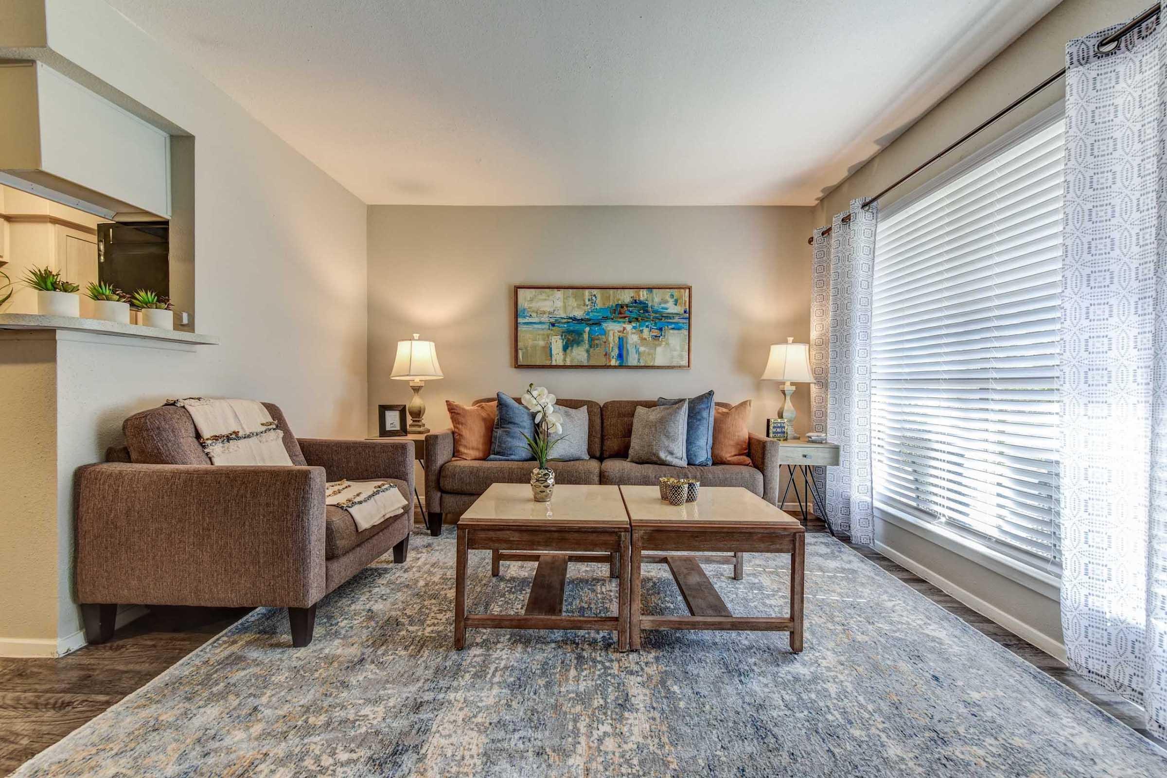 Cozy living room with a brown couch and armchair, light-colored walls, and large windows covered with sheer curtains. A wooden coffee table sits on a decorative rug, and a modern painting hangs above the couch. Two lamps provide ambient lighting for a warm atmosphere.