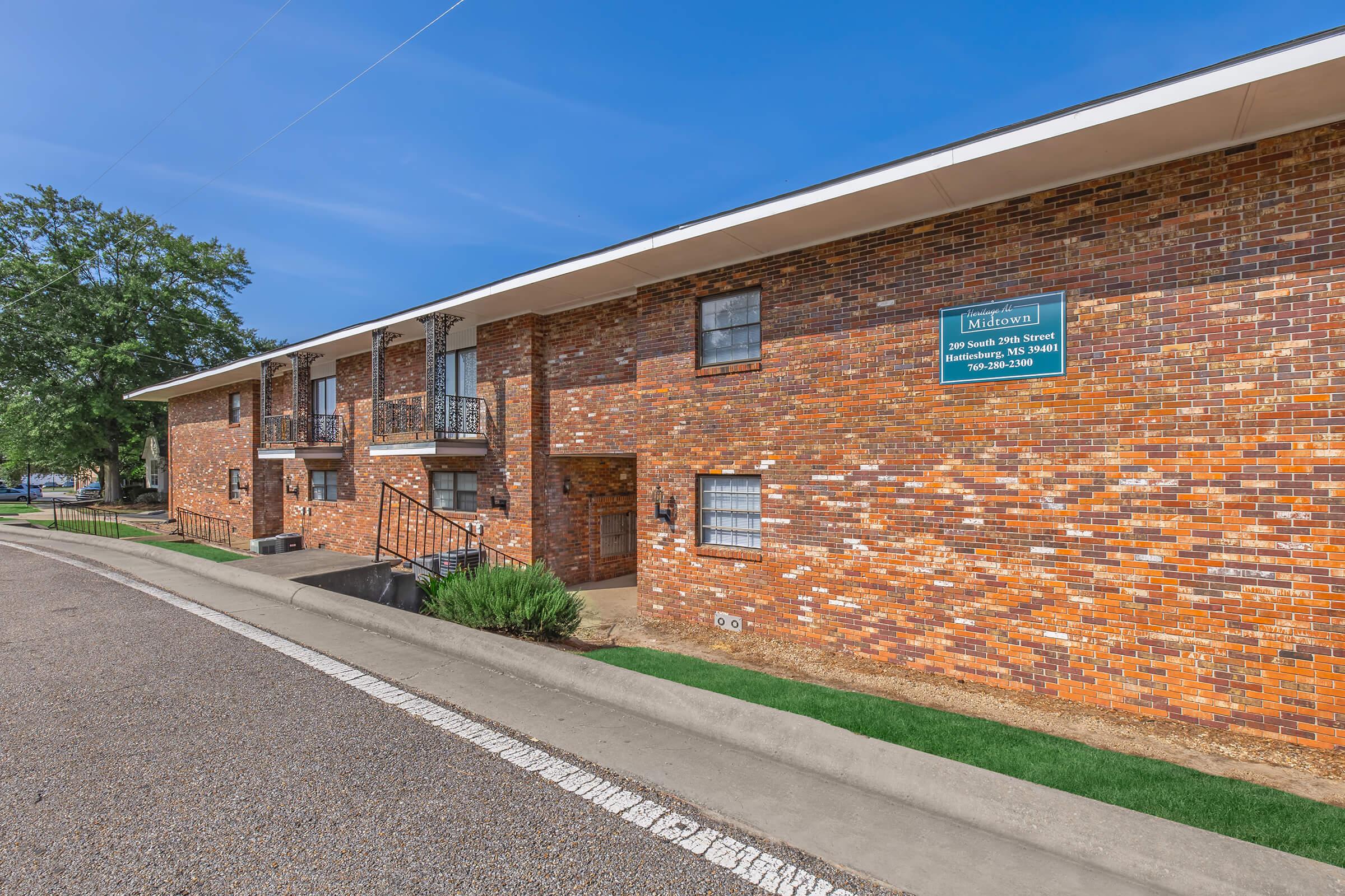 Two-story brick apartment building with a well-maintained exterior. The building features multiple windows and a sign indicating the property name and contact information. The surrounding area has grassy patches and a paved road leading up to the entrance. Clear blue sky above.