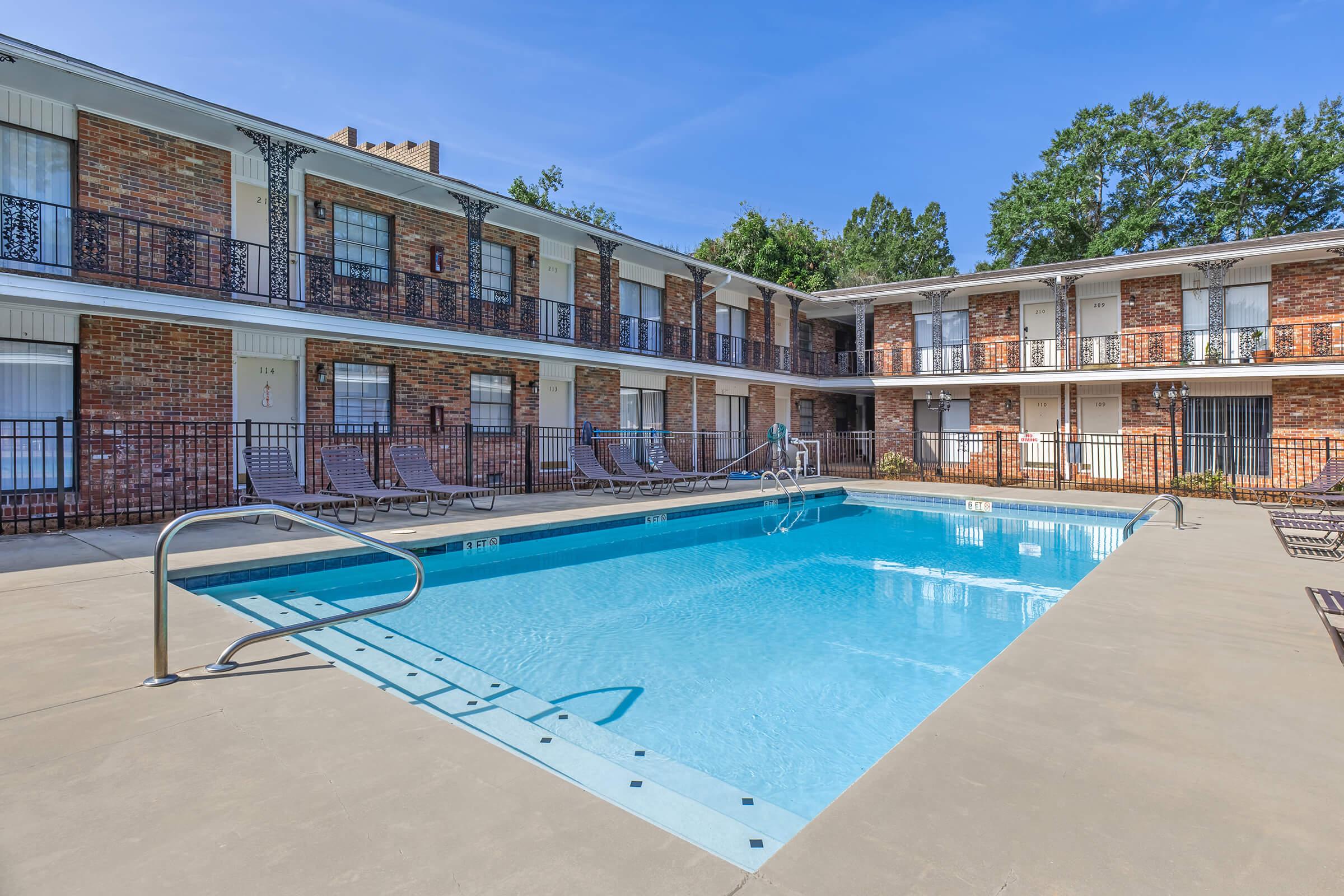A clear blue swimming pool surrounded by a patio area with lounge chairs. The pool is located in the center of a two-story brick building with balconies and several windows. Lush greenery is visible in the background under a sunny sky.