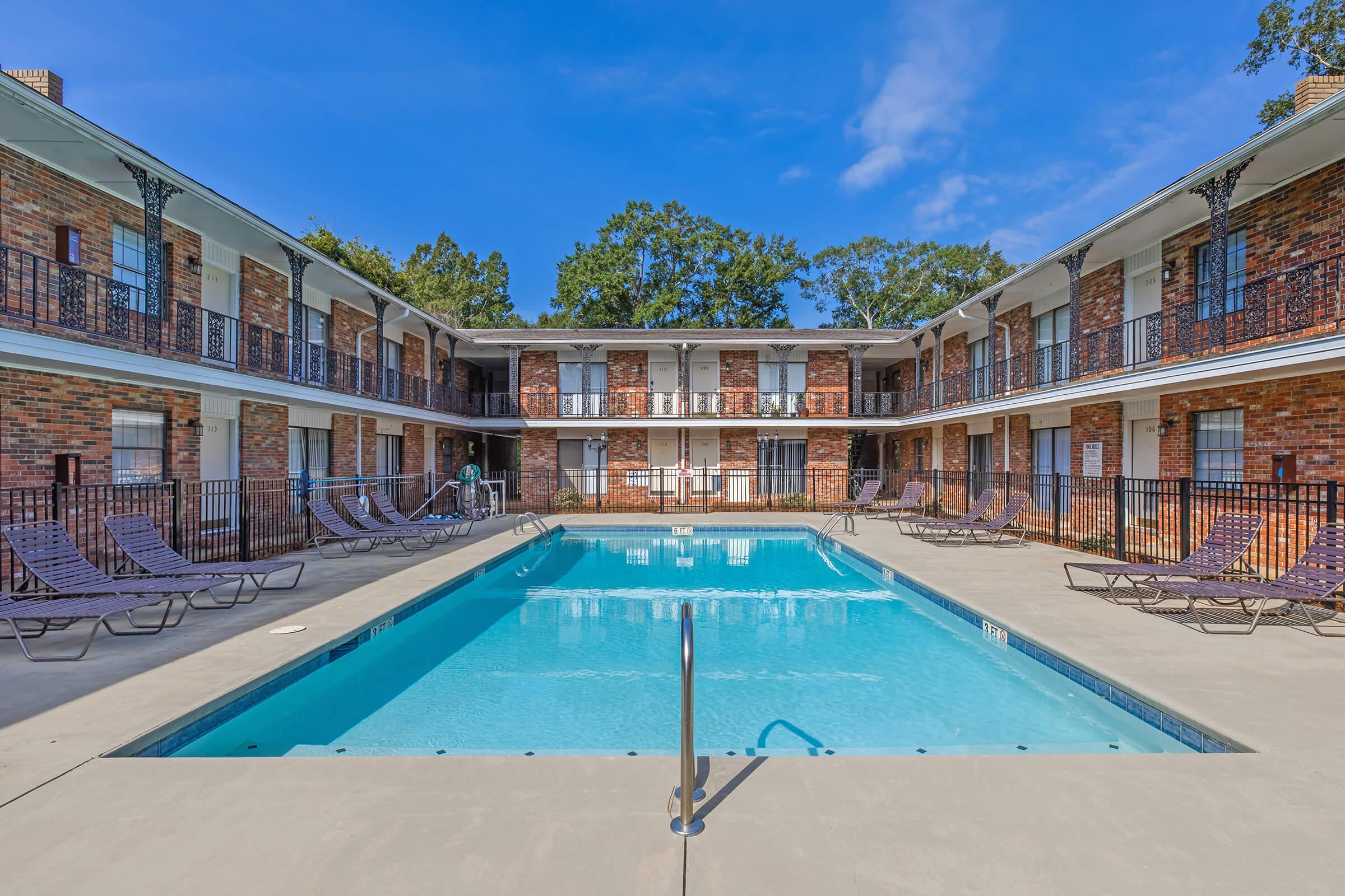 A clear blue swimming pool surrounded by a courtyard with brick buildings. Loungers are placed around the pool area, and trees are visible in the background under a bright blue sky. The setting appears inviting and well-maintained, ideal for relaxation or social gatherings.