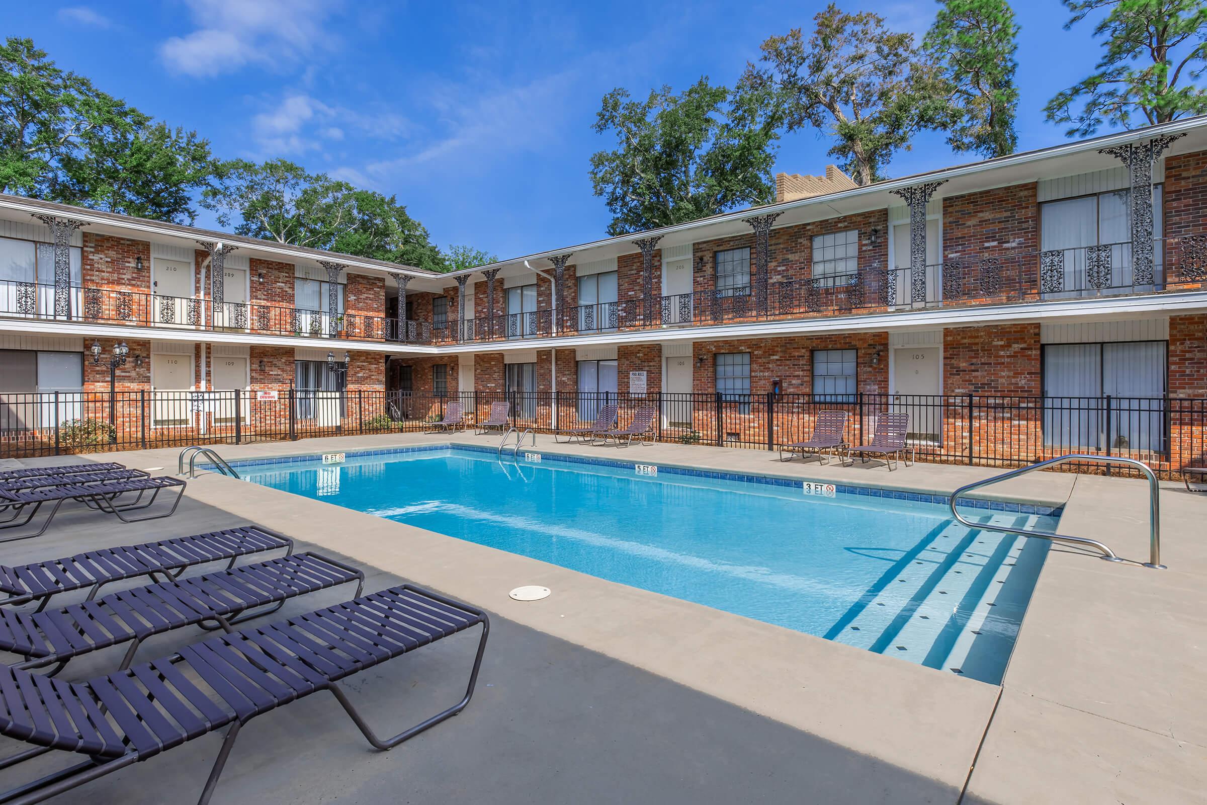 A sunny courtyard with a rectangular pool surrounded by lounge chairs. Two levels of brick apartment buildings are visible in the background, featuring balconies with railings. Lush green trees and a bright blue sky add to the inviting atmosphere.