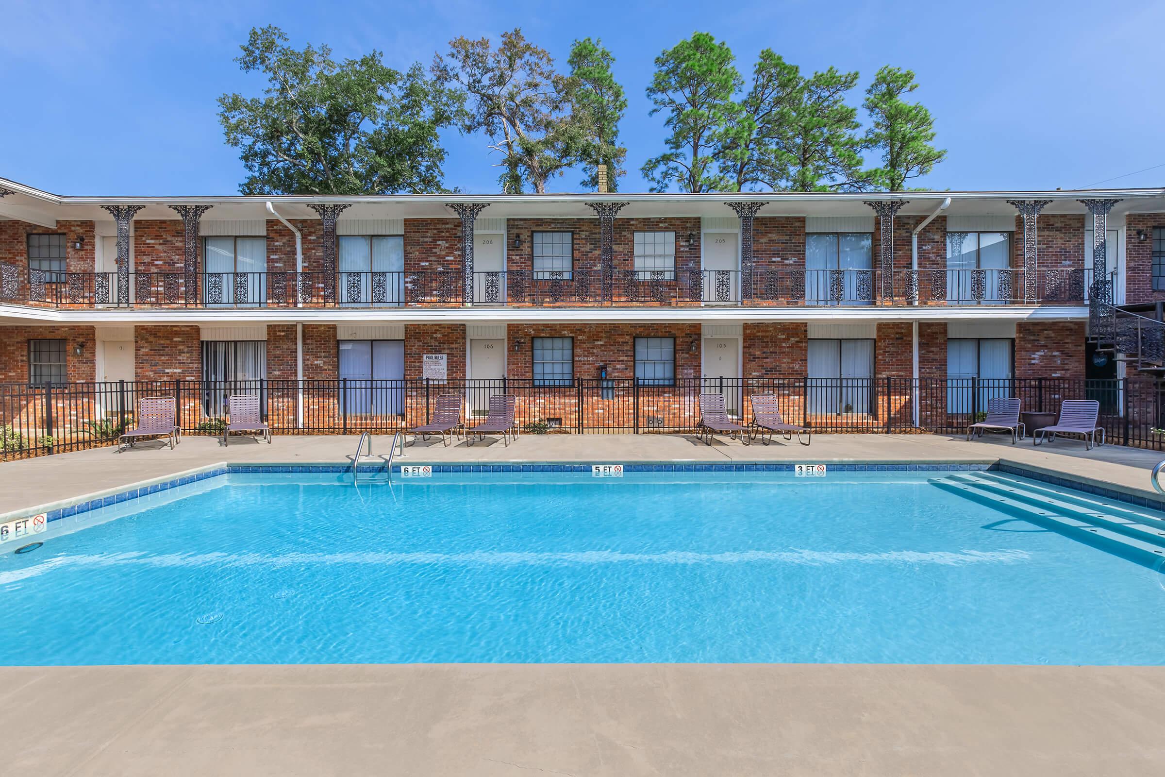 A clear blue swimming pool in the foreground with sun loungers on the deck. In the background, a two-story brick building with balconies overlooks the pool. Lush green trees are visible above the building against a clear blue sky.