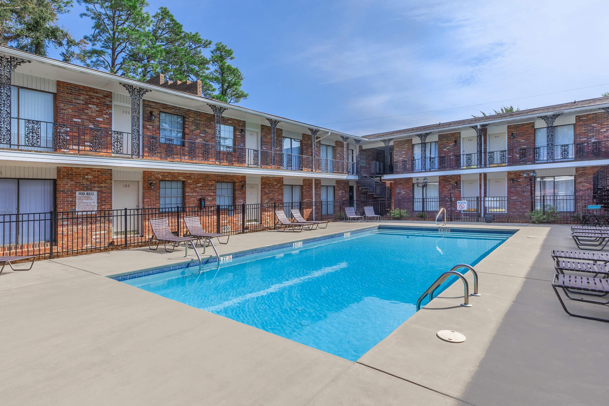 A clear, inviting swimming pool located in the center of a brick apartment complex. Surrounding the pool are lounge chairs, with several trees in the background providing shade. The sky is partly cloudy, indicating a pleasant day. The atmosphere is relaxed and welcoming.