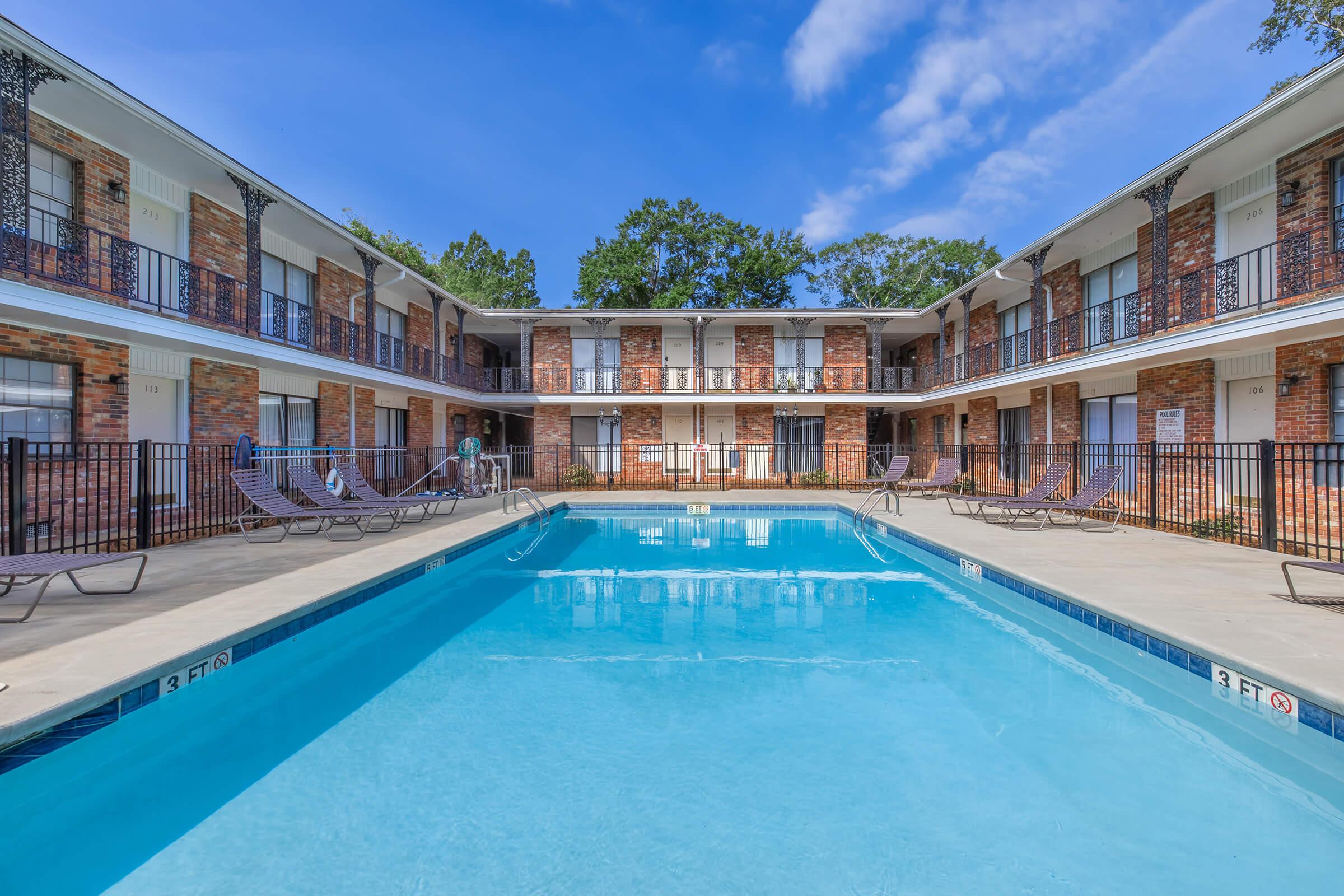 A clear blue swimming pool surrounded by a brick apartment complex. Lounge chairs are arranged near the pool, and the building features multiple balconies overlooking the pool area. The sky is bright with scattered clouds, highlighting a sunny day.