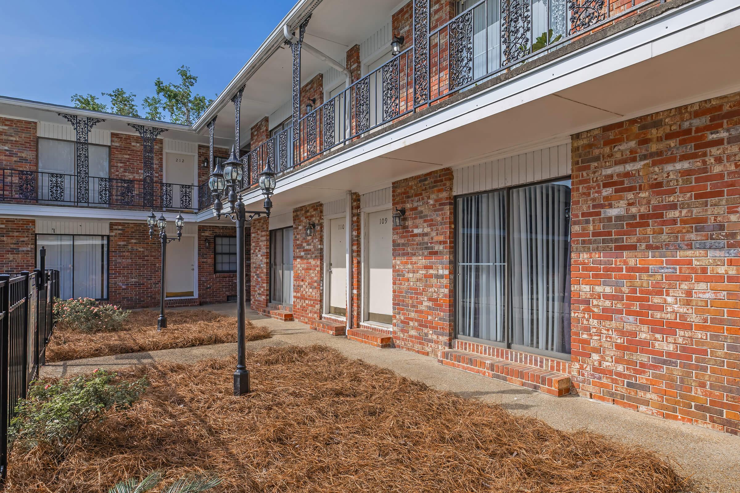 A charming view of a two-story brick apartment building with a walkway lined with pine straw, featuring multiple doors and decorative wrought-iron balconies. Street lamps add a quaint touch to the outdoor area, surrounded by greenery. The setting is bright, showcasing clear blue skies.