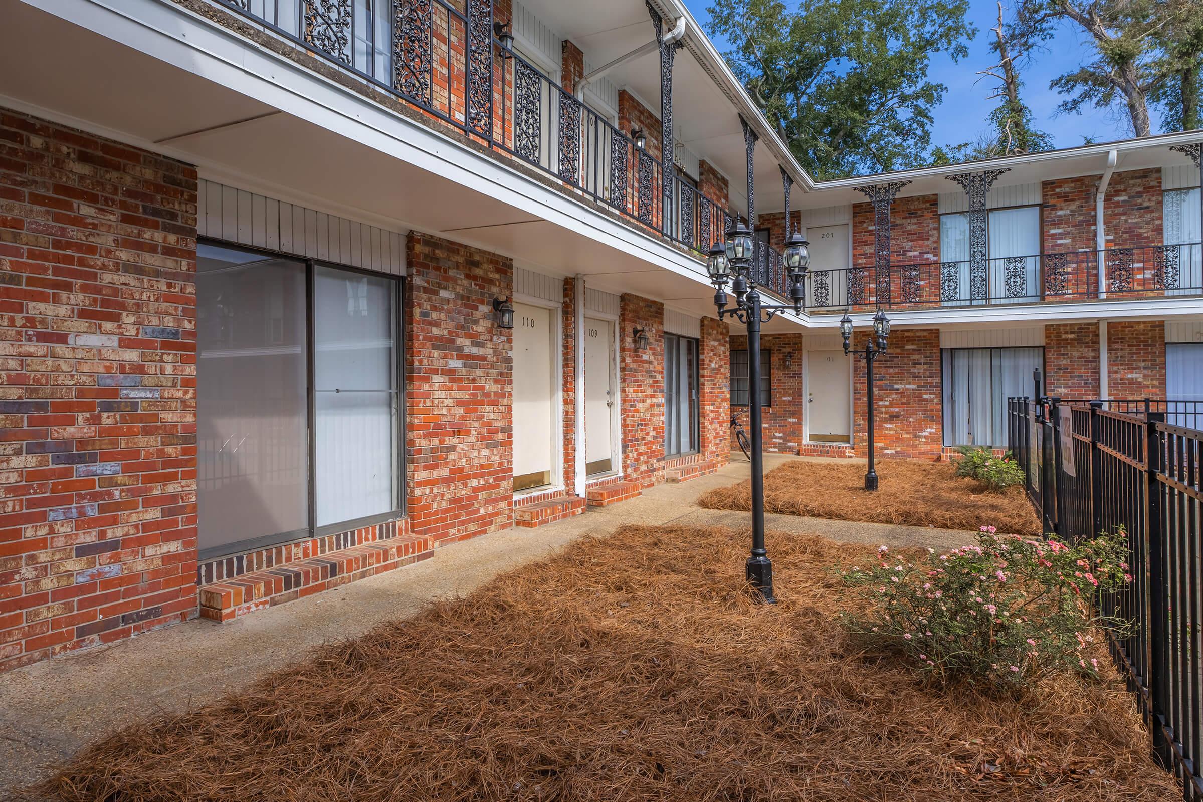 Exterior view of a brick apartment building featuring multiple floors with balconies. There are walkways leading to individual units, surrounded by landscaped areas with mulch and small plants. Decorative lampposts line the walkways, and trees are visible in the background under a clear blue sky.