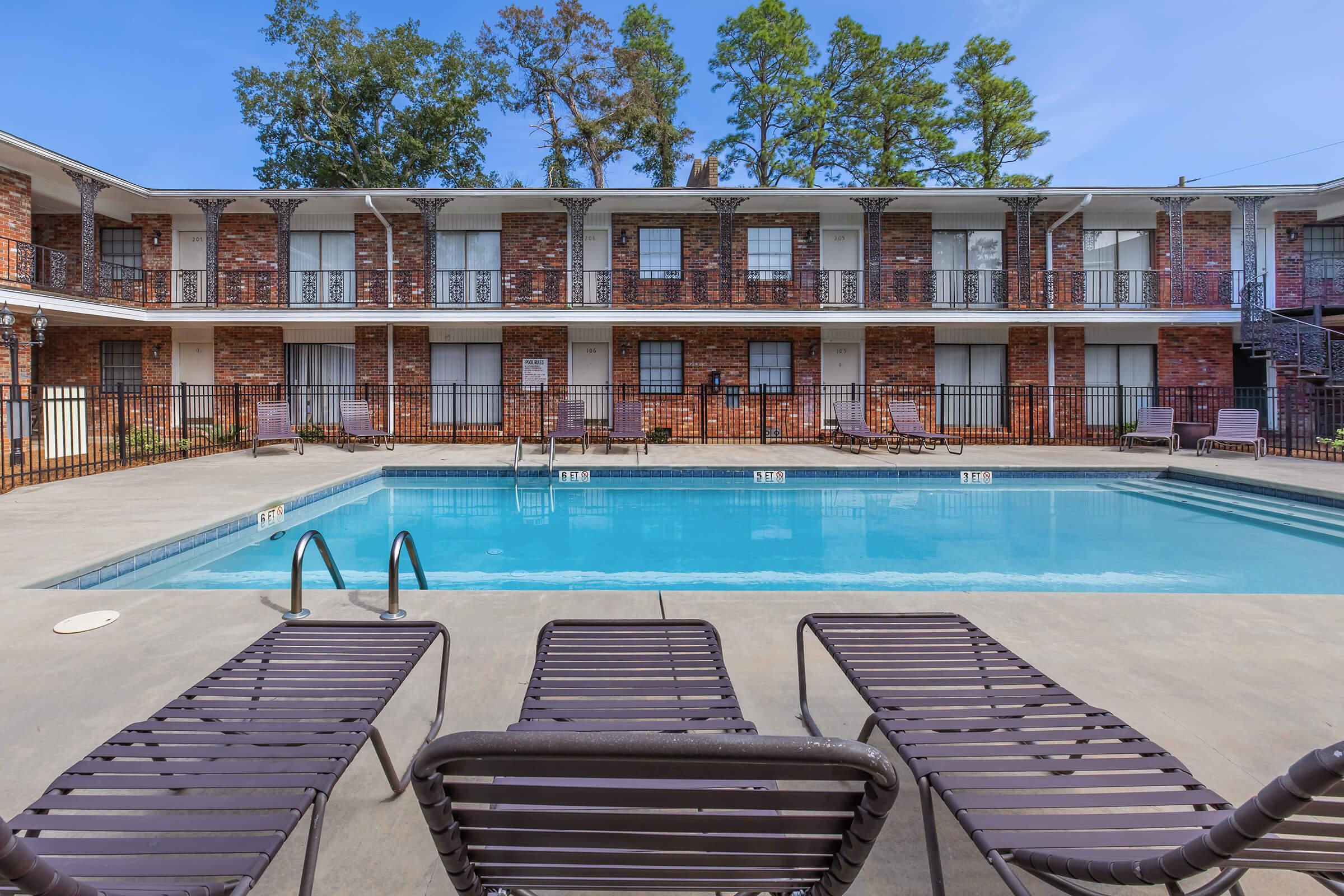 A swimming pool surrounded by lounge chairs at an apartment complex. The pool area features clear water, and the building in the background has brick exteriors with balconies. Trees are visible in the background, suggesting a sunny outdoor environment.